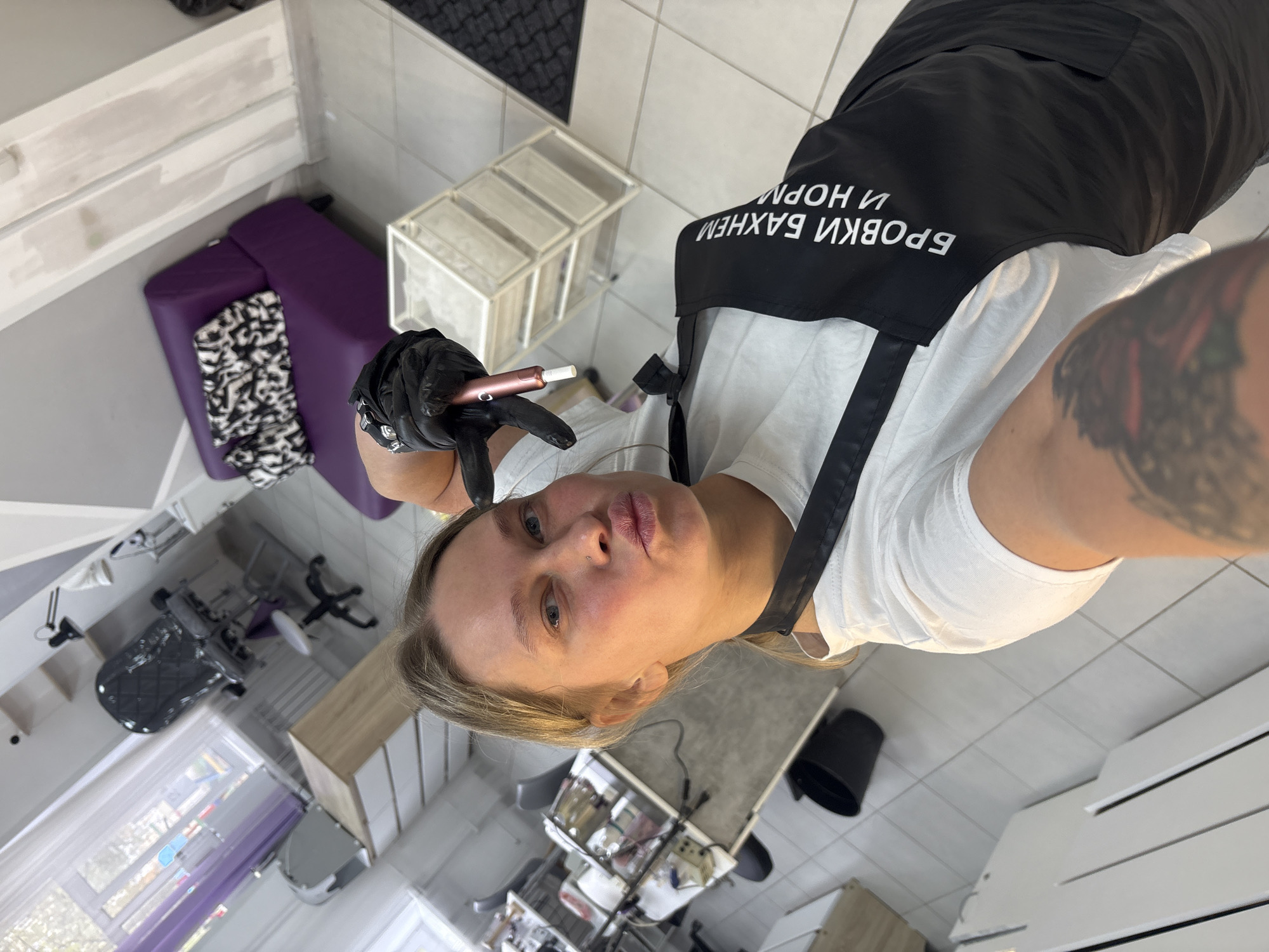 A woman with blonde hair is taking a selfie in what appears to be a beauty salon. She is wearing a white t-shirt and a black apron with the text "БРОВКИ В АХНЕМ И НОРМ" in Russian. She is wearing a black glove on her left hand and is holding a pink device with a white tip. The background includes a purple beauty bed with a black and white zebra-print cover, a white storage unit, and a tiled floor. A tattoo is visible on her right arm. This is most likely located in Perm, Russia, given the Russian text on the apron.