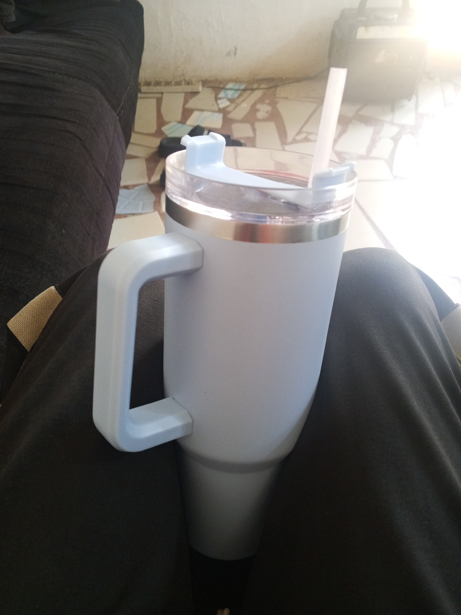 A light blue, insulated tumbler with a handle and a clear plastic straw sits on a person's lap. The person is wearing dark-colored pants. Part of a dark gray couch is visible on the left. The background shows a tiled surface and a glimpse of an object, possibly a bag, in a brightly lit area. The location is unknown.