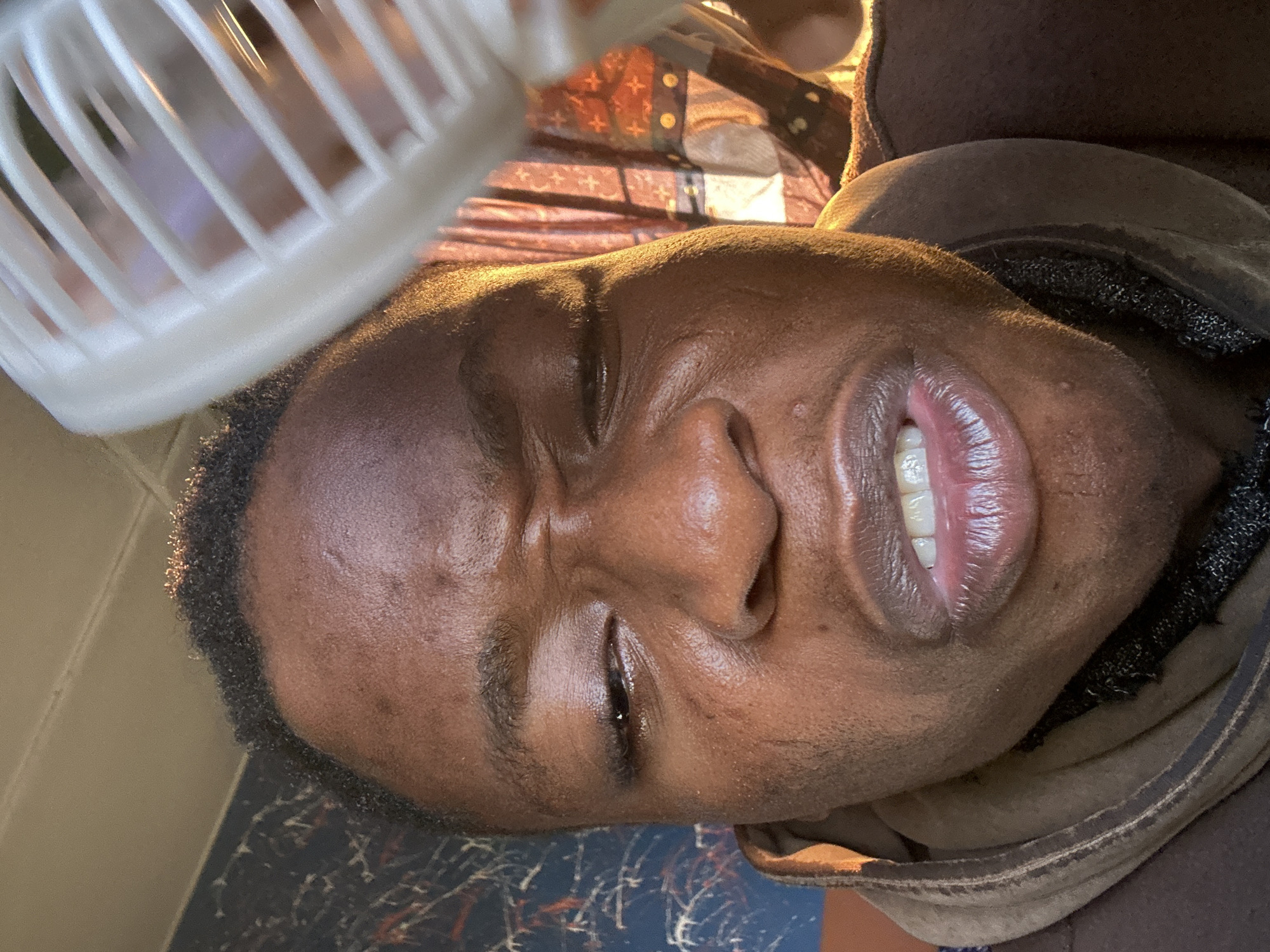 A close-up, tilted shot shows a person with dark skin and short, curly hair, their eyes closed. A small white plastic fan is positioned above their head. The person wears a brown, hooded garment. The background is blurred, revealing a patterned surface. The location is unknown.