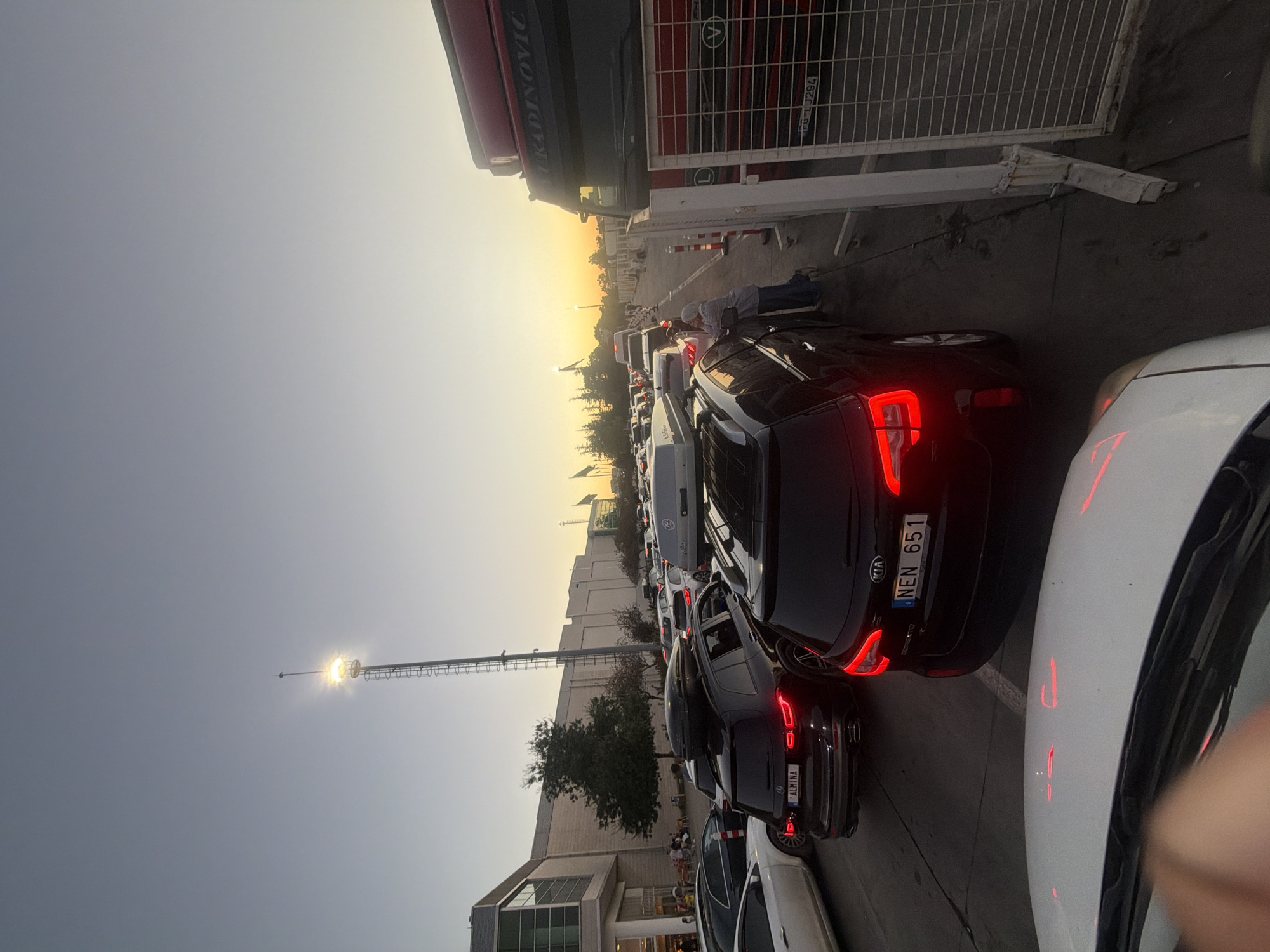 The image depicts a traffic queue at what appears to be a border crossing or toll plaza in Edirne Merkez, Turkey, during sunset. The sky is a gradient of blue and orange, indicating the sun is low on the horizon. In the foreground, the hood of a white car is visible, with red reflections on its surface.
Behind the white car, a black Kia Sorento is stopped, displaying a license plate that reads "NEN 651". To its left is another black SUV, possibly a Mercedes-Benz, with a license plate that reads "ALMINA". Both SUVs have roof boxes, suggesting travel. Further back, a long line of vehicles is visible, including cars and possibly some larger trucks.
On the right side of the frame, a red truck with "TRADINOVIC" written on its side is parked. Adjacent to the truck is a metal fence with a grid pattern. A person wearing a headscarf is standing next to one of the cars, looking towards the traffic.
In the background, a tall tower with a bright light illuminates the scene. Buildings, trees, and flagpoles are also visible, consistent with a commercial or logistical hub. The overall impression is of a busy, potentially slow-moving situation at a transportation point.