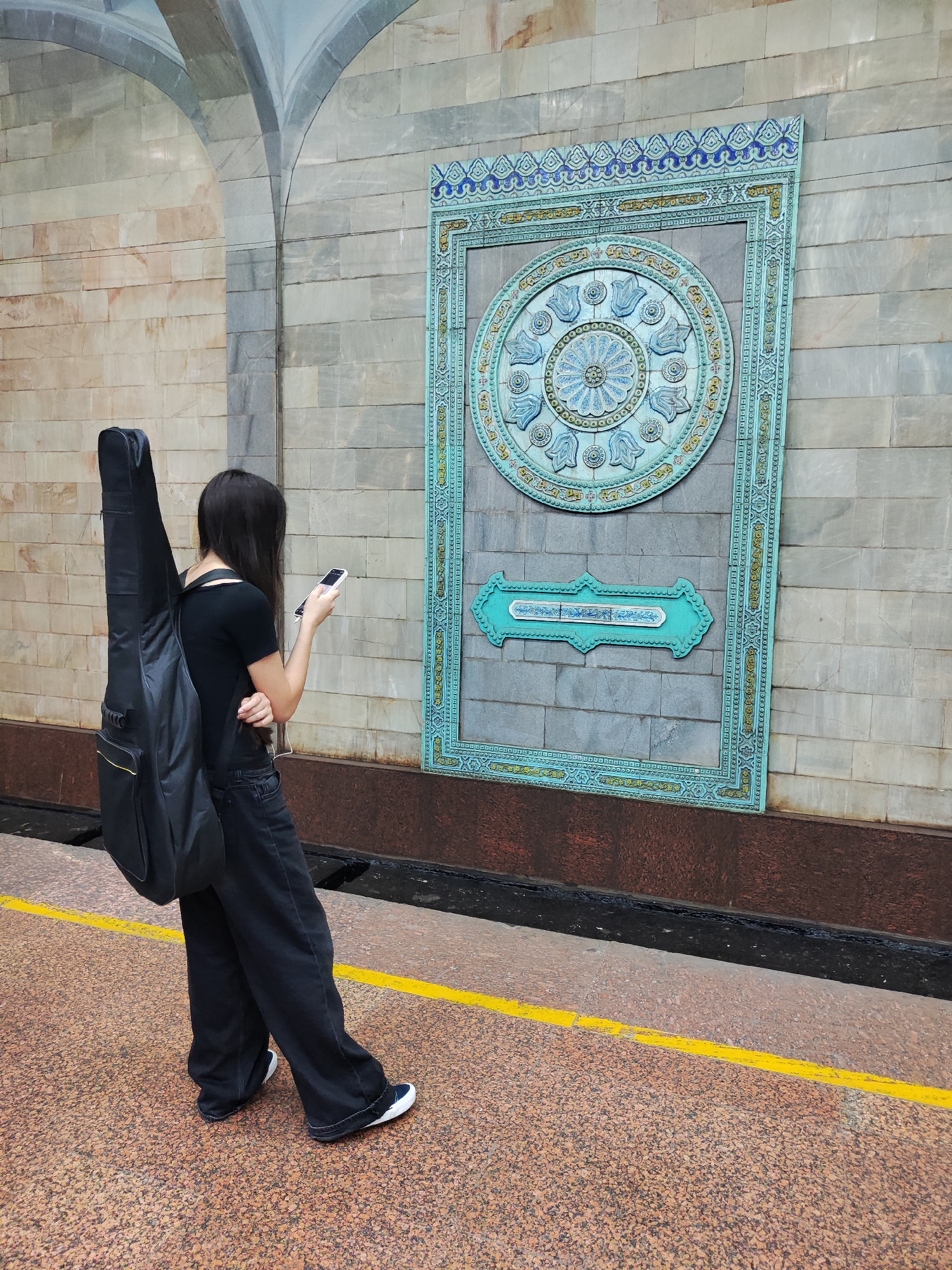 A person is standing on a platform with a yellow line, holding a smartphone and wearing a black t-shirt, black wide-leg pants, and black and white sneakers. A black guitar case is slung over their back. The background features a tiled wall with an ornate, turquoise and blue decorative mosaic panel featuring floral and geometric patterns. The architectural style suggests a metro station in Tashkent, Uzbekistan, possibly at the Kosmonavtlar metro station, known for its elaborate artistic decorations.