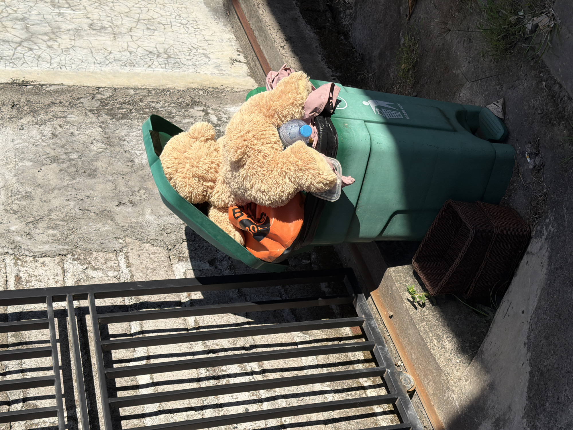 A green trash receptacle with its lid open is located outside, near a concrete structure. Inside the trash receptacle are various items including a large teddy bear and a water bottle. To the side is a dark brown wicker basket. A section of a black metal gate can be seen in the lower left corner. The location appears to be Kuta Selatan, Indonesia.