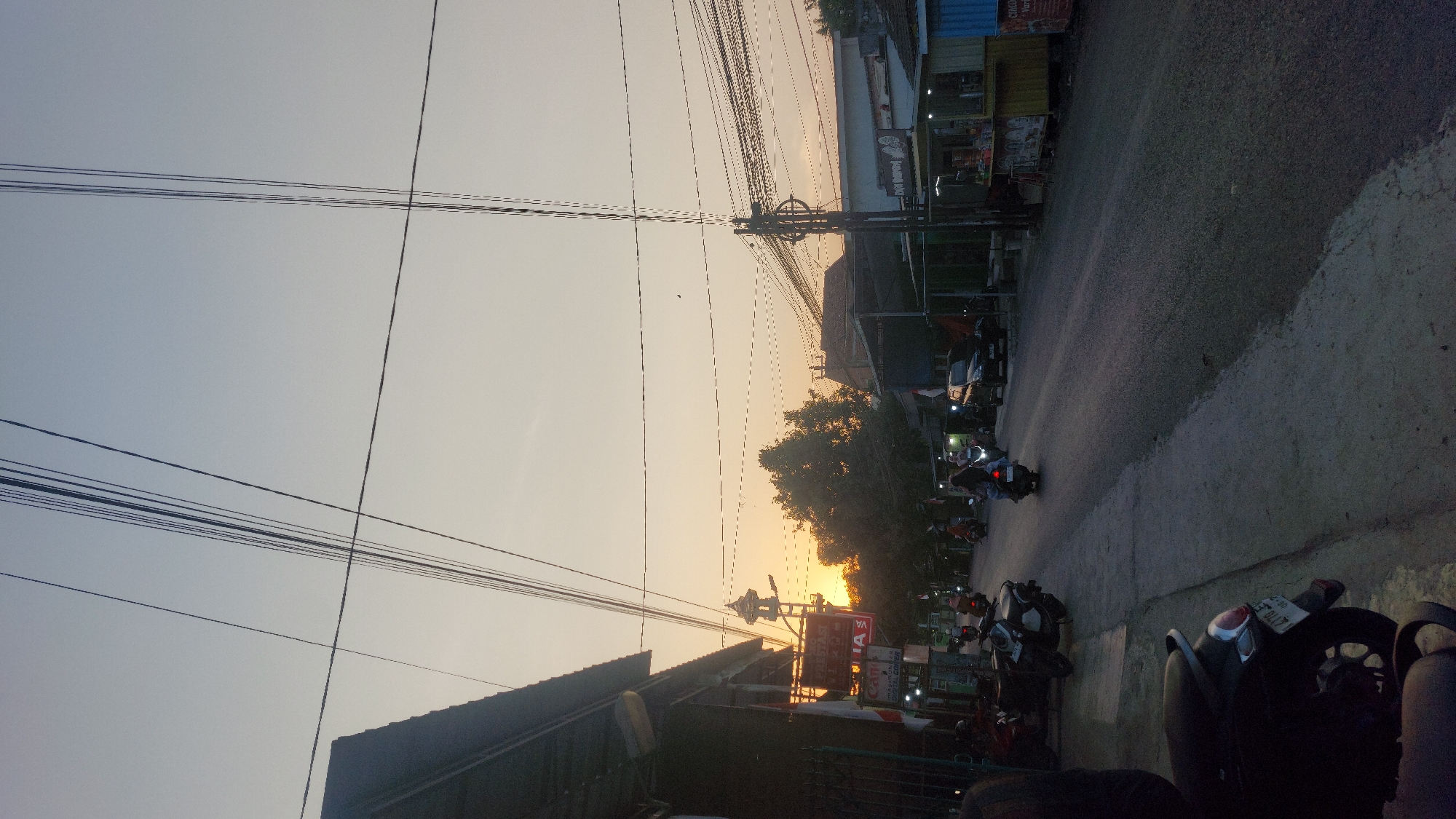This is a medium shot, captured at street level, of a street in the city of Karas, Indonesia, during what appears to be either sunrise or sunset. The sky is a soft gradient of pale orange and light blue, indicating a low sun position. Numerous overhead electrical wires crisscross the frame, casting thin shadows on the sky.
On the left side of the street, a low-lying building with a dark roof and a metal fence is visible. A sign with red lettering is partially obscured. Further down the street, a tall, slender structure resembling a minaret or tower is visible against the bright sky, with trees surrounding its base.
The street itself is paved with asphalt and appears to be moderately trafficked by motorcycles and a few cars. Several motorcycles are parked on the left side of the road, and others are being ridden. Some individuals are riding the motorcycles, most of whom are wearing helmets and some are wearing headscarves. On the right side of the street, there are several small food stalls or kiosks with brightly colored awnings. One stall has a yellow base and a blue awning, while another to its right has a red and white awning. Signs in a local script are visible on the stalls. The overall atmosphere is that of a bustling but quiet urban environment during a transitional period of the day.