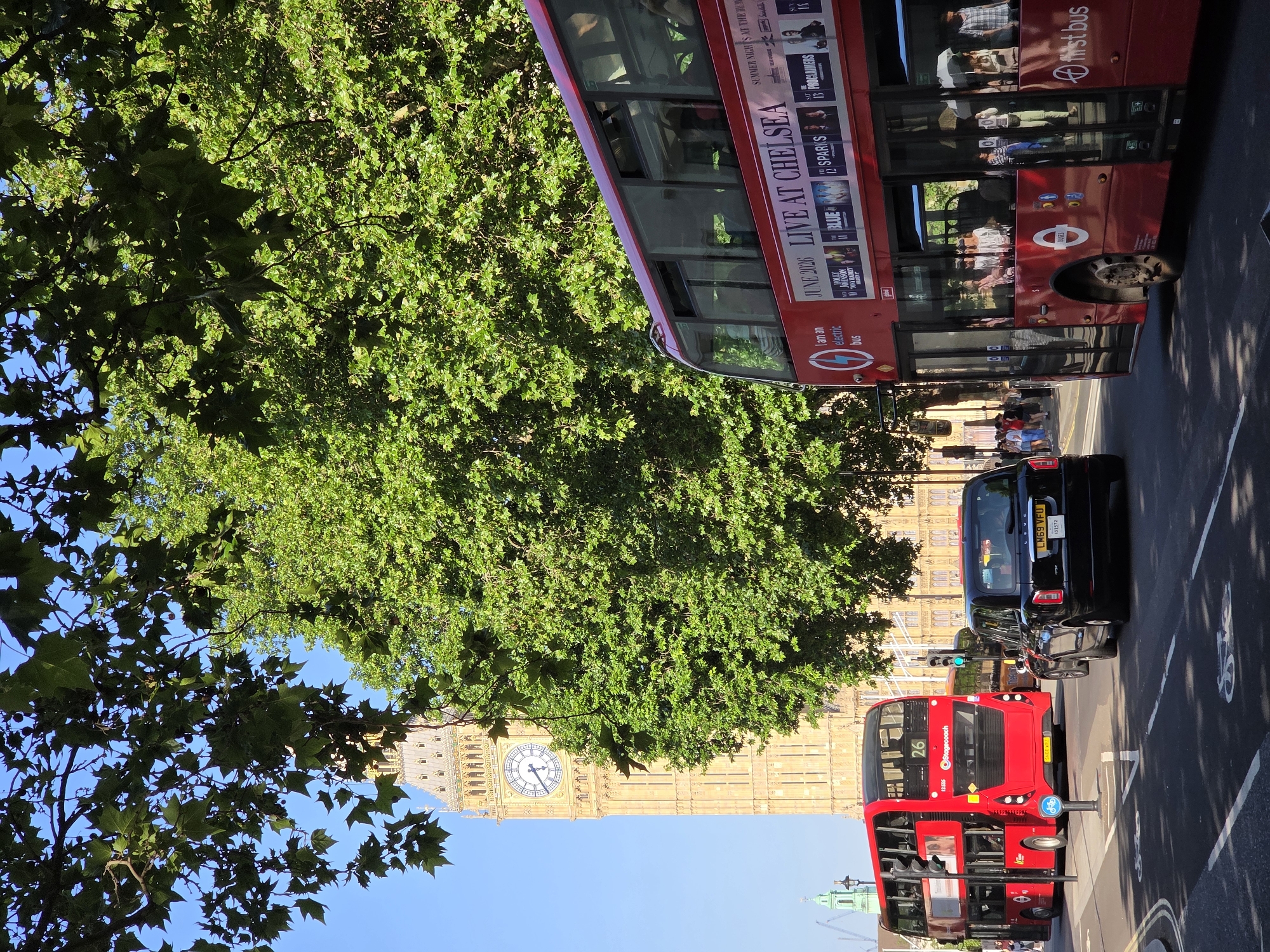 The image depicts a street scene in London, United Kingdom, featuring a partial view of the Elizabeth Tower (Big Ben) and the Palace of Westminster, seen through and around foliage. Two red double-decker buses are visible, one positioned closer to the camera, labeled as an electric bus, with the advertisement "LIVE AT CHELSEA JUNE 2026". A black taxi cab is situated in the middle of the street, and pedestrians can be seen in the background. Street markings for bike lanes are also visible.