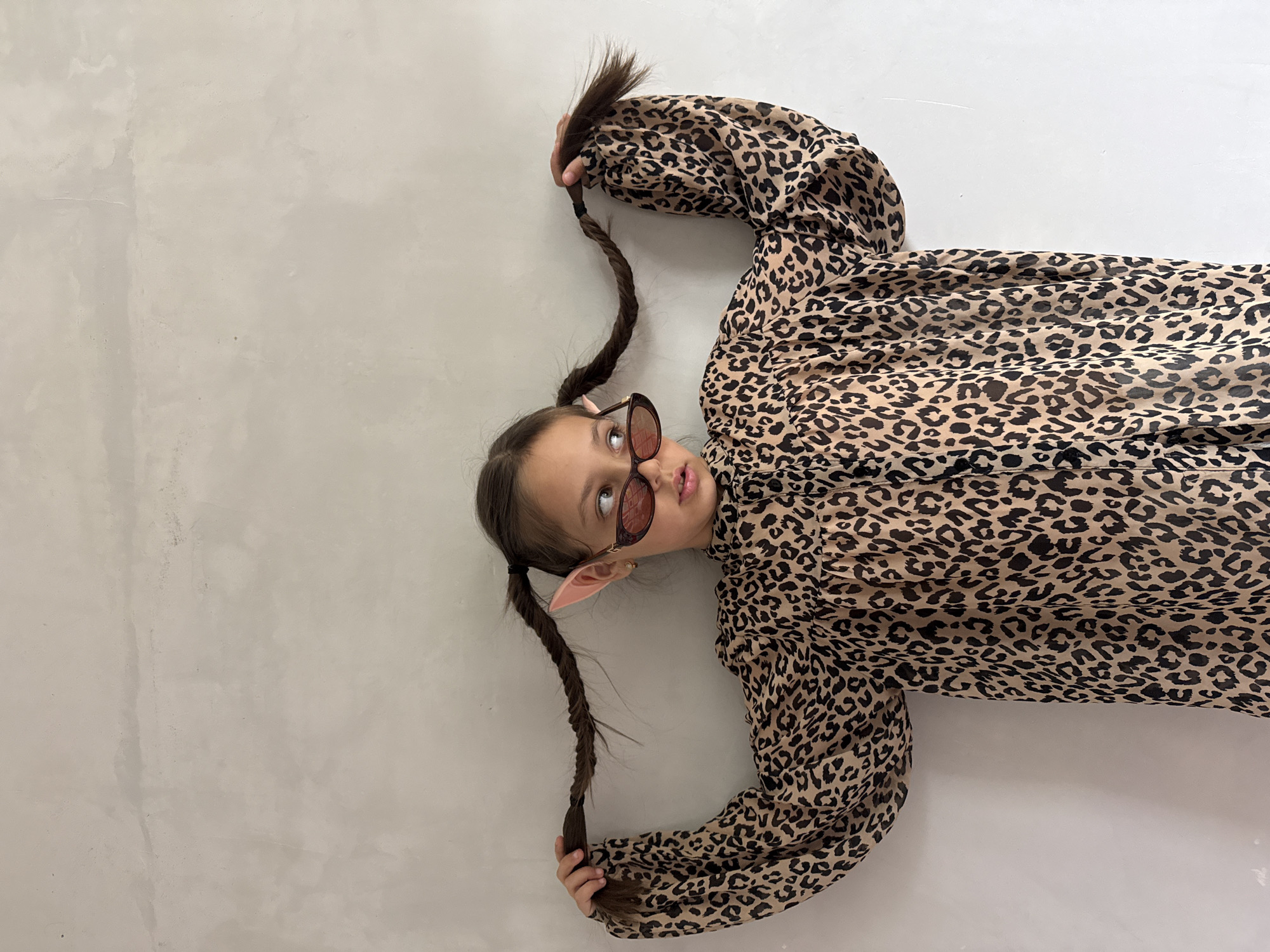 A young girl lies on a light gray surface. She has brown hair styled into pigtails with pink pointed ears, and she's wearing pink sunglasses. She's dressed in a leopard-print top with long, puffy sleeves and is holding the ends of her pigtails. The location is unknown, but the city is specified as Votkinsk, Russia.