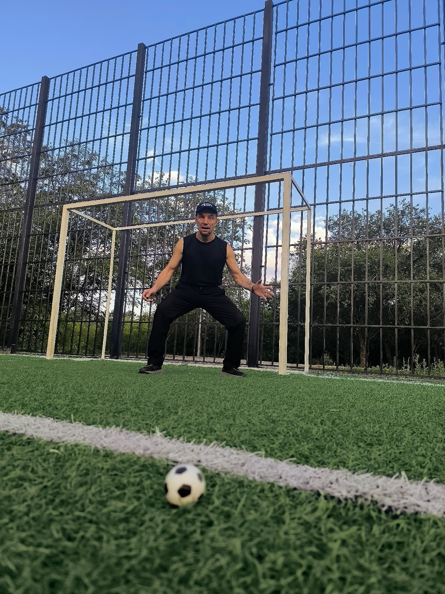 A man in a black sleeveless shirt and black pants stands on a soccer field in a defensive stance, with his arms outstretched. A small soccer ball is visible in the foreground, positioned in front of the man. A soccer goal is behind him, and a tall metal fence encloses the field. The background consists of trees and a blue sky with some clouds. The image appears to be taken outdoors, possibly in a park or sports facility. The location is specified as the city of Qarağandı, Kazakhstan.