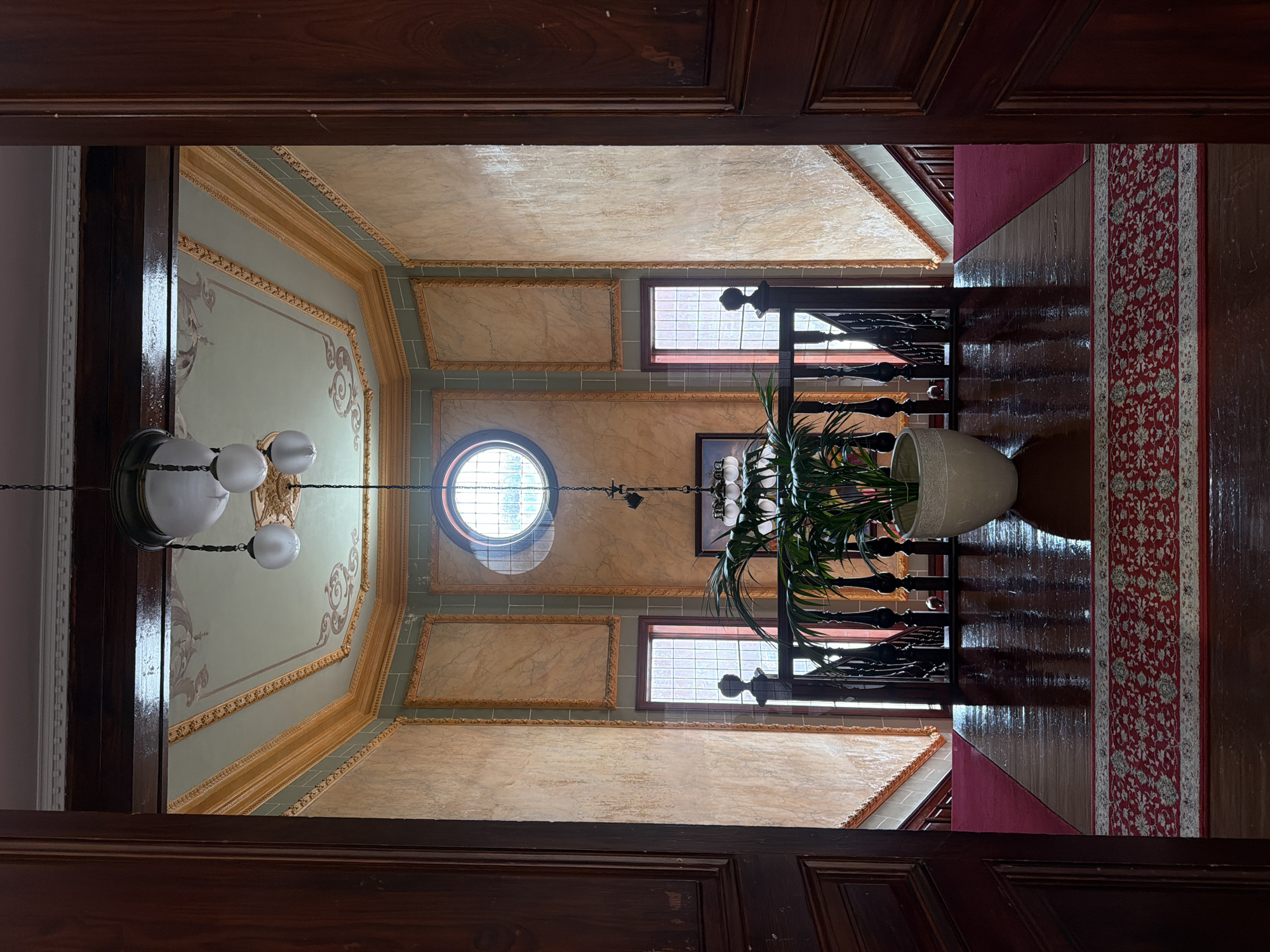 The image depicts the interior of a grand staircase landing within a historic building in La Orotava, Spain. The perspective is from an open doorway, looking into a brightly lit, opulent space. The landing features highly polished dark wood floors, partially covered by a deep red carpet with an intricate pattern. To the left and right, dark wooden banisters with turned spindles ascend the stairs, which are also carpeted in red.
The walls are adorned with textured panels in shades of peach and cream, framed by ornate gold moldings. Above, a decorative ceiling is painted with classical motifs in muted tones of green and gold. A prominent feature in the center of the landing is a large, potted green plant with broad leaves, placed in a textured white ceramic pot.
Dominating the upper portion of the scene is a circular window with a grid pattern, allowing natural light to filter into the space. Suspended from the ceiling by chains is a vintage light fixture with three spherical glass globes. The overall impression is one of refined elegance and historical preservation.