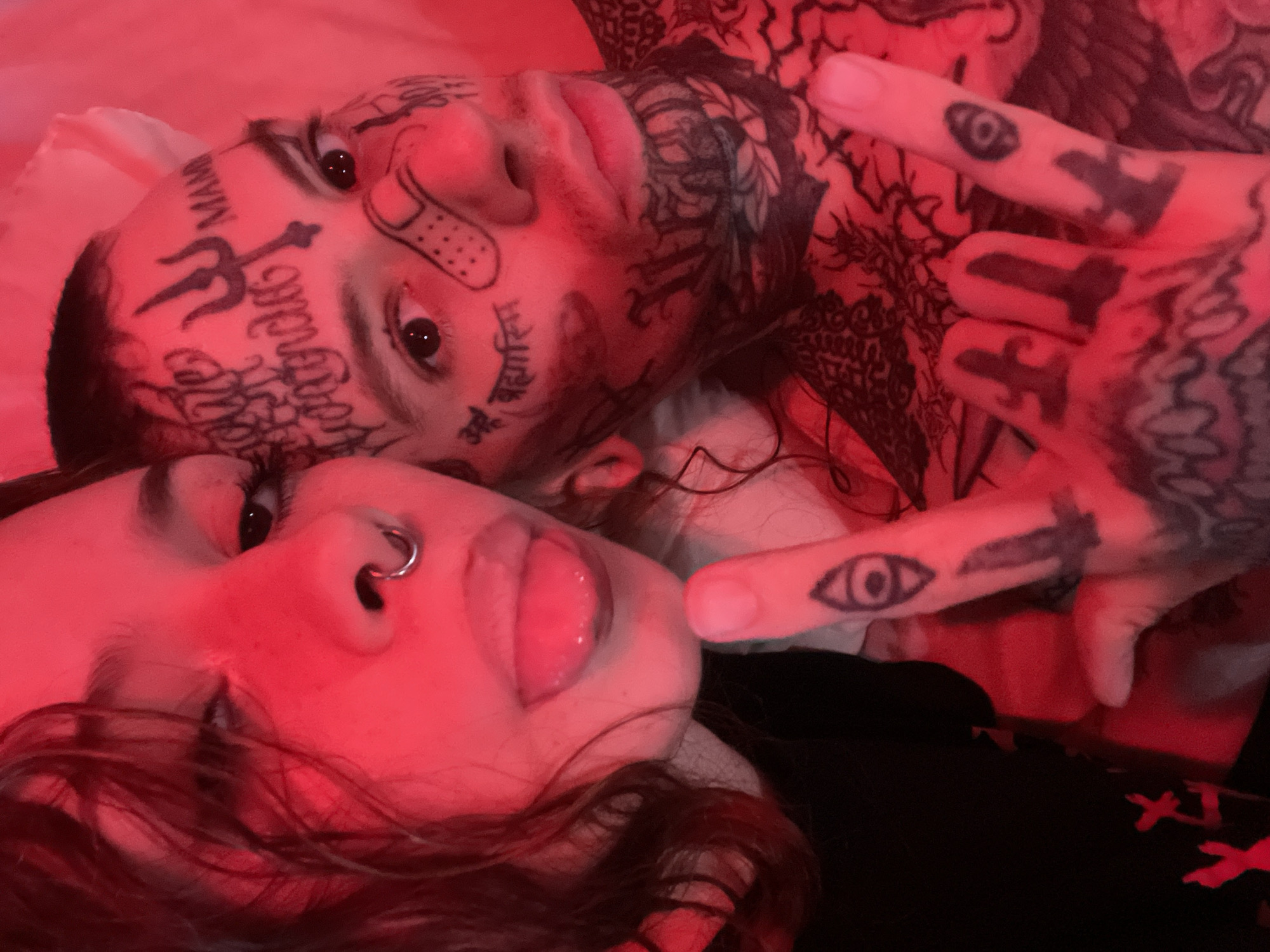 A close-up, intimate photograph depicts two individuals bathed in a pervasive red light, suggesting a nocturnal or mood-lit setting, possibly in Buenos Aires, Argentina. The camera angle is from slightly above, with both subjects lying down or reclined.
In the foreground, a person with dark, wavy hair is visible, primarily showing their face from the nose down. They have a silver septum piercing and their tongue is slightly extended, giving a playful or expressive look.
Behind and slightly above, a second individual is prominently featured, heavily tattooed across their face, neck, chest, and hands. Distinct facial tattoos include a black "Trishul" (trident) symbol between their eyebrows, the word "MAMA" above their right eyebrow, and script on their left cheek which appears to be in Devanagari, possibly reading "ॐ बकासुरम" (Om Bakasuram). A bandage-like drawing is also visible on the right cheek. Their neck and chest are covered with intricate dark patterns. This person is making a hand gesture with fingers pointed upwards, and an "all-seeing eye" tattoo is visible on one of their fingers, alongside other symbolic hand tattoos.
Both individuals are in close proximity, suggesting a personal connection. The entire scene is drenched in a striking red hue, which highlights their features and tattoos, creating an intense and passionate ambiance. The background is indistinct and soft, likely indicating a bed or plush surface.