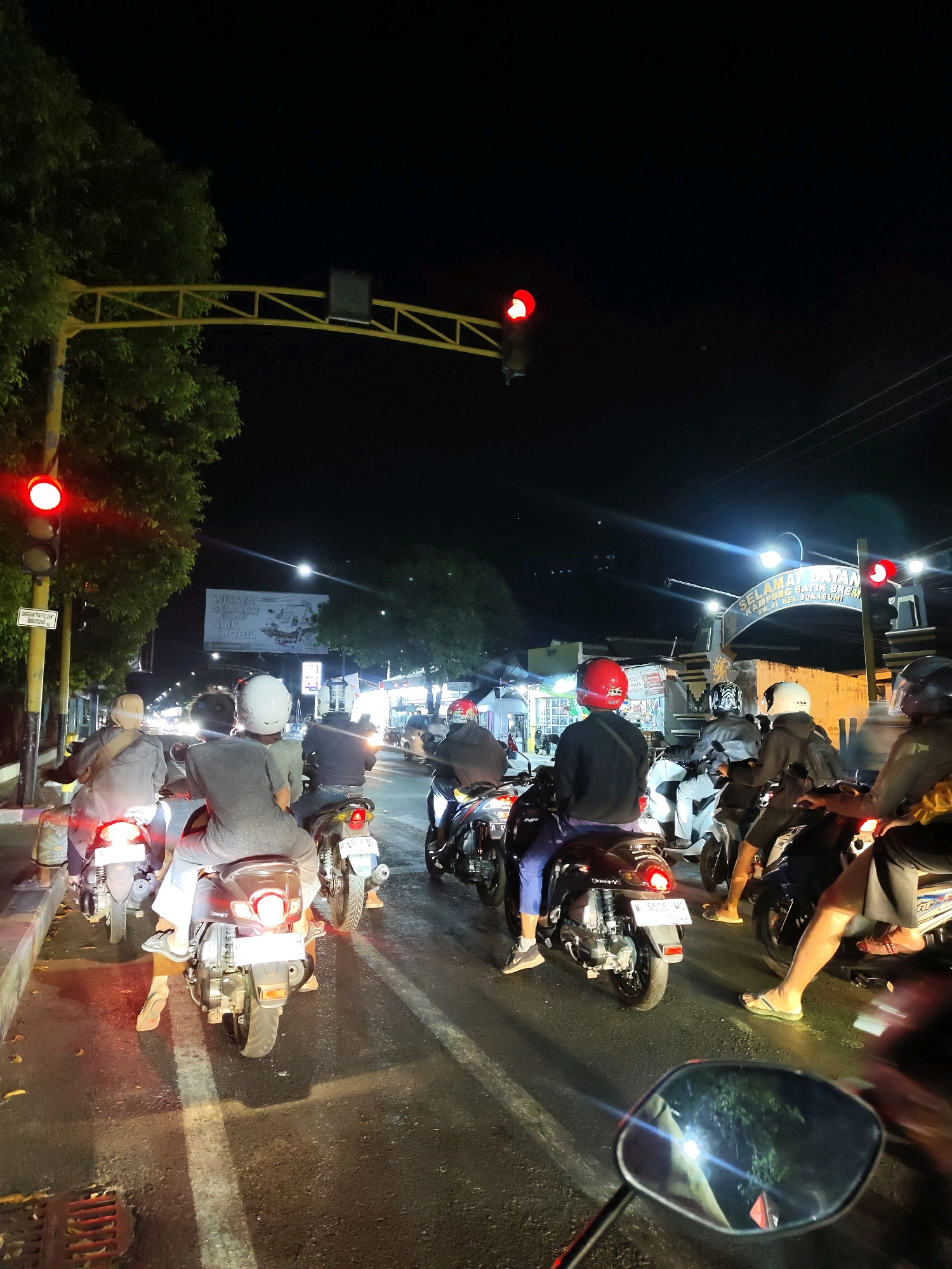 The image depicts a traffic scene at night in Probolinggo, Indonesia. A group of motorcyclists are stopped at a traffic light, which is red. The road is wet, suggesting it may have recently rained. Trees line the left side of the road, and a yellow traffic signal gantry spans overhead. Several motorcycles are visible, with riders wearing helmets of various colors, including white and red. Some riders are wearing casual clothing, such as t-shirts and shorts. In the background, there are streetlights, buildings, and a billboard. The overall atmosphere is that of a typical urban traffic situation at night.