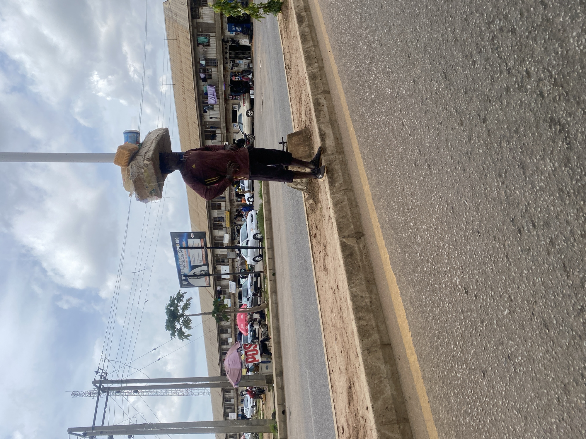 A man stands on a curb on the side of a road in New Stadium, Nigeria, balancing a large, flat package topped with two containers on his head. He is wearing a maroon long-sleeved shirt with yellow accents and dark shorts, and black athletic shoes. Behind him, a multi-story building with shops on the ground floor stretches across the background. Laundry is hung from the balconies of the upper floors. Cars are parked and driving on the road, and a pink umbrella and a smaller red umbrella are visible near a stall on the left side of the frame. The sky is cloudy.