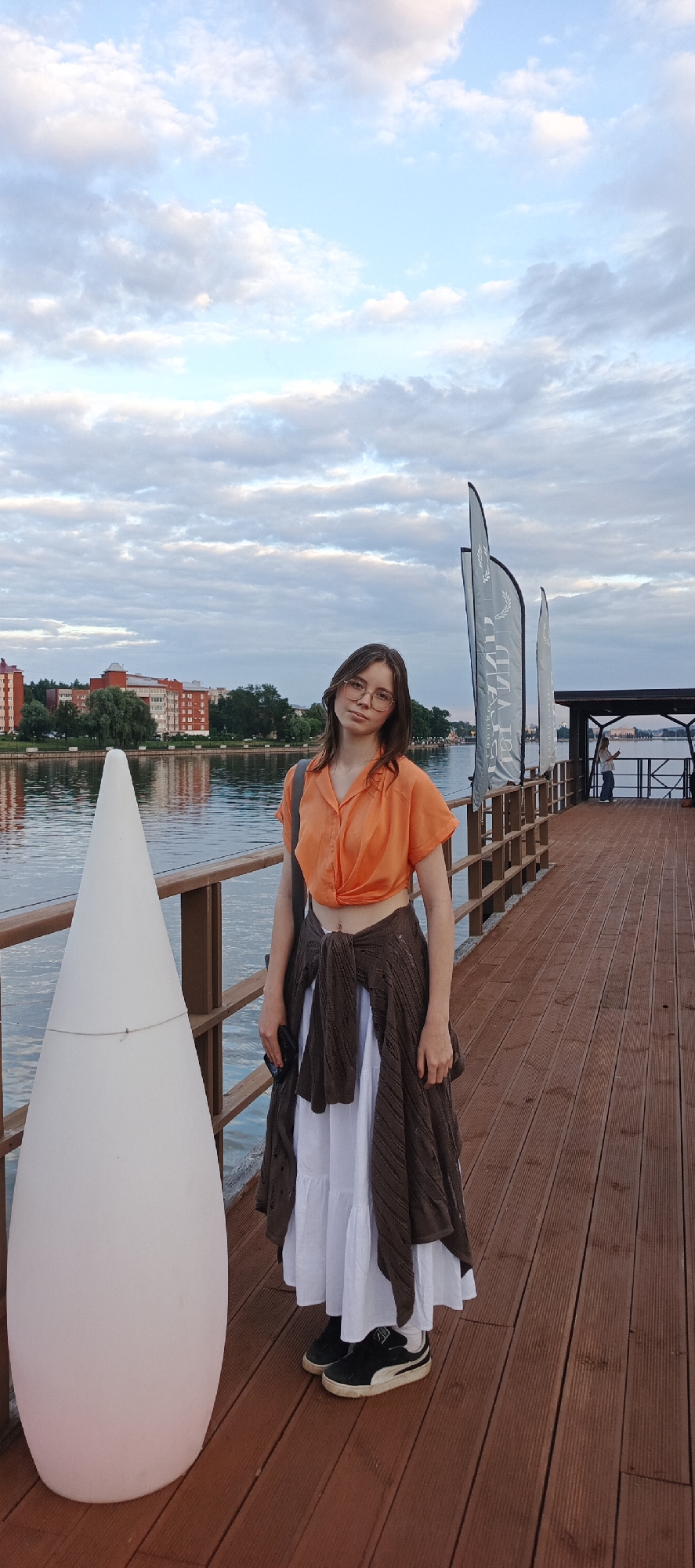A young woman with brown hair, wearing glasses, an orange cropped blouse, a white tiered maxi skirt, and a brown cardigan tied around her waist, stands on a wooden pier. She is looking towards the camera. To her left is a large white conical lamp. In the background, a calm body of water reflects a cloudy sky and buildings on the opposite bank, suggesting the location is Votkinsk, Russia. The pier has a railing and several tall flags.