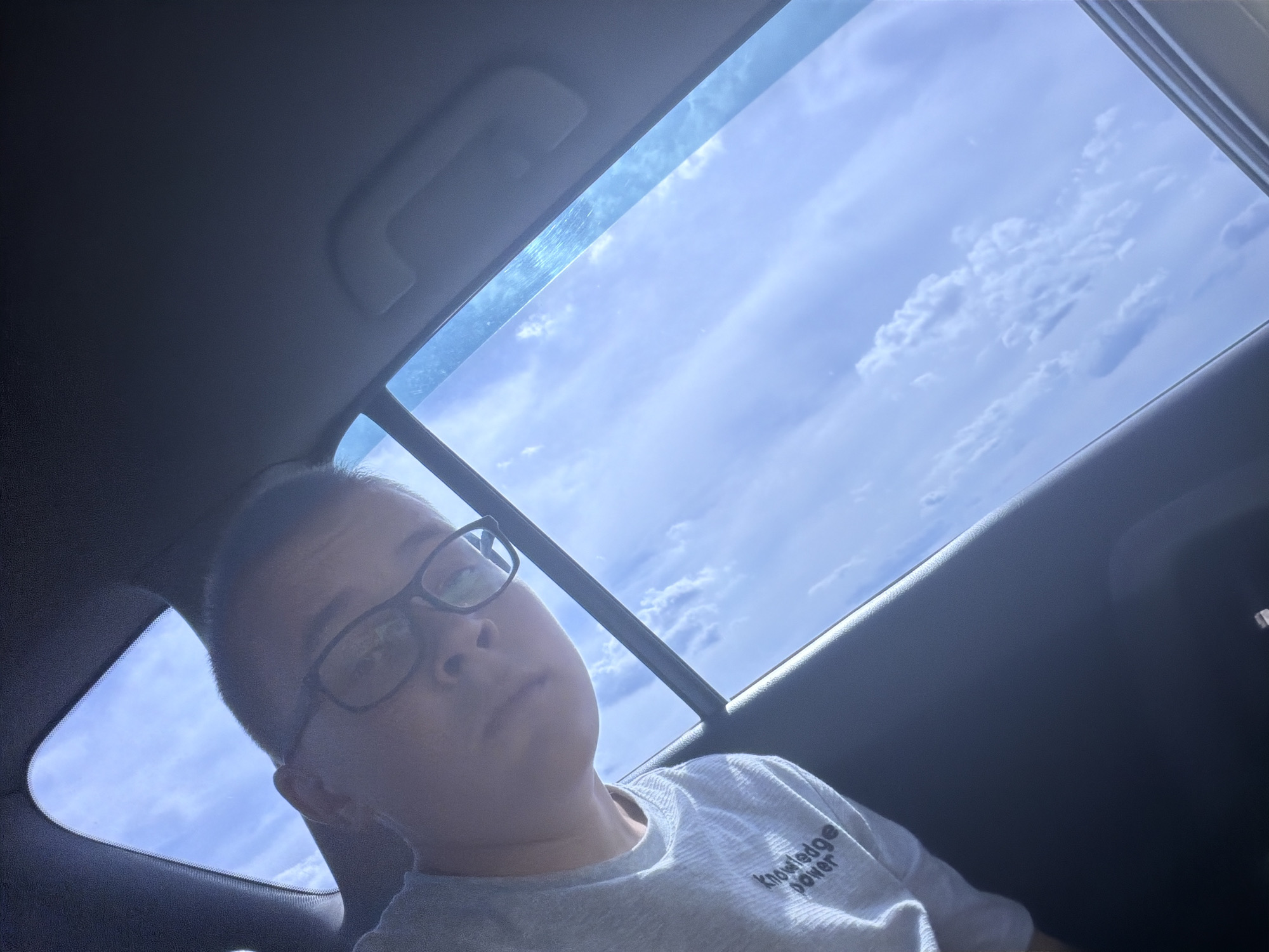 The original image is a low-angle, selfie-style photograph taken inside a vehicle, looking up towards the sky through the sunroof and side window. The subject, a person wearing glasses and a light gray t-shirt with the text "knowledge power" printed on it, is positioned in the foreground, partially visible on the left side of the frame. The person's face, with short dark hair and glasses, is turned towards the camera.

The background displays a clear blue sky with scattered white clouds, suggesting daylight and fair weather. The car's interior is dark, with the sunroof and side window frames creating geometric shapes within the frame. The overall impression is that of a person capturing a moment while traveling in a car on a sunny day.

This image was taken on July 16, 2023, at approximately 12:43 PM local time, in the village of Novoe Teneevo, Russia. The coordinates are 55.1932 degrees North latitude and 49.3162 degrees East longitude. The ambient temperature was around 25°C (77°F), with a light breeze from the west.