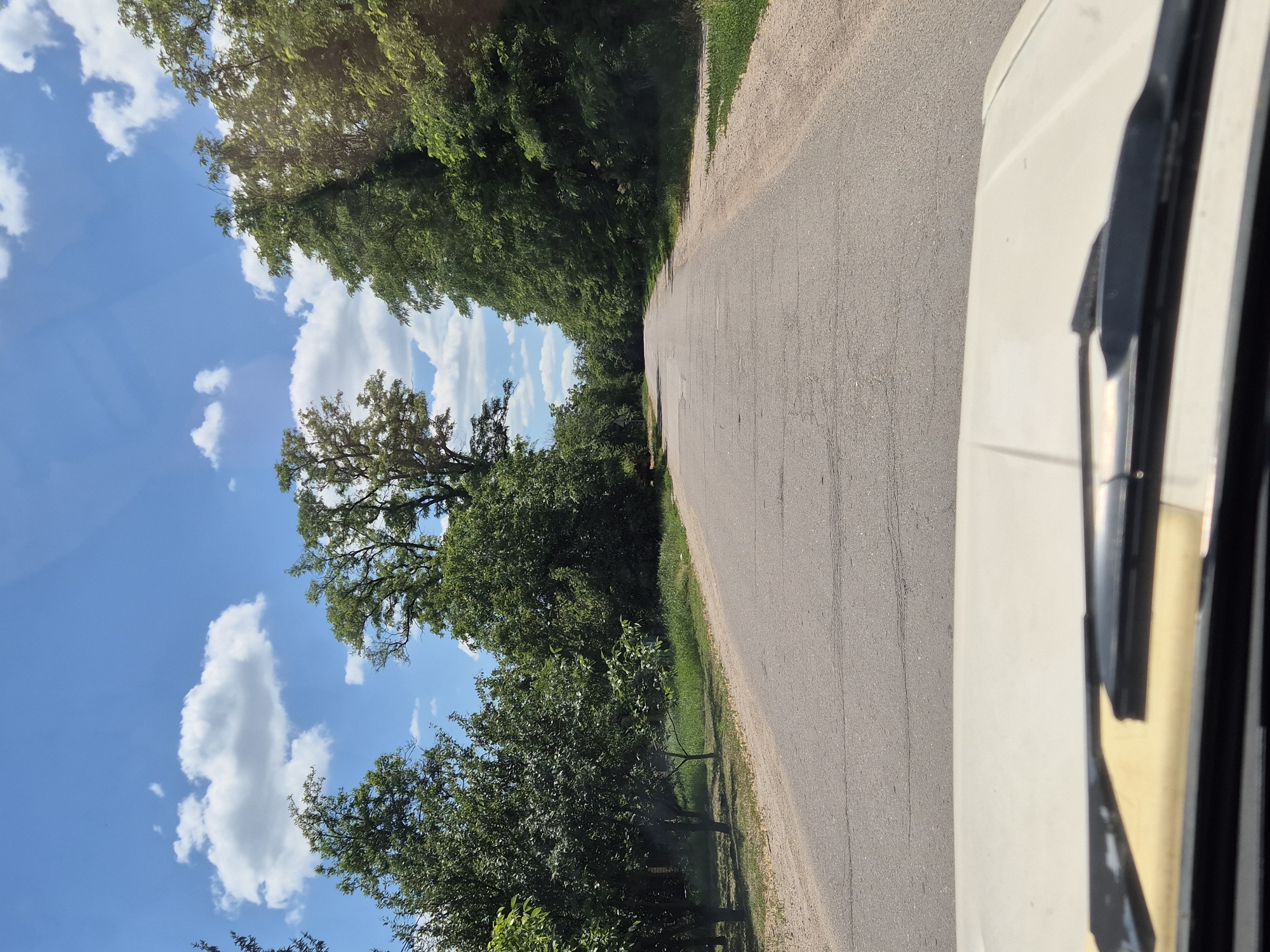 A view from inside a vehicle traveling on an asphalt road, which is lined with trees on both sides. The road curves to the left in the distance. The sky is blue with scattered white clouds. The foreground shows the hood and windshield wipers of the vehicle. The environment suggests a rural or suburban setting, possibly in Sozonivka, Ukraine, given the presence of deciduous trees and the overall natural landscape. There are no people visible in the image.