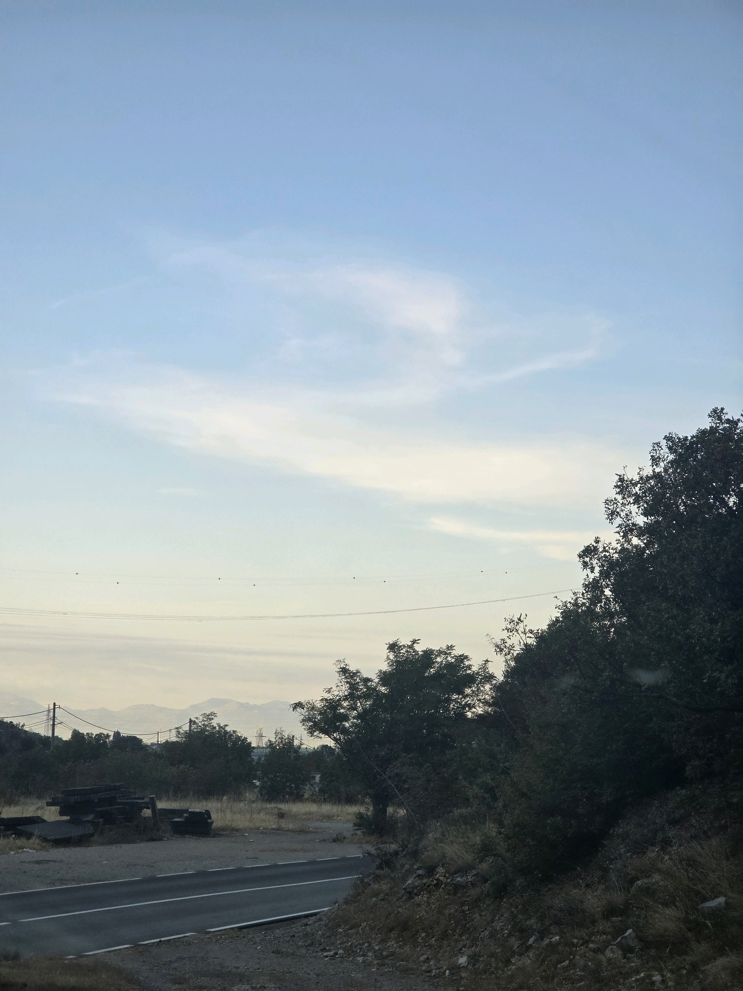 The image depicts a rural roadside scene in the outskirts of Podgorica, Montenegro. A paved road with a white line runs through the foreground, bordered by dry grass and rocky terrain. To the left, a pile of dark, rectangular objects, possibly discarded construction materials or large stones, sits next to the road. Beyond the road, a dense cluster of green trees and bushes forms a natural barrier. In the distance, silhouetted against a pale blue sky with wispy clouds, are rolling hills or mountains. Power lines with a few small, dark objects (likely insulators or birds) are visible strung across the middle ground, connecting to utility poles. The overall impression is one of a quiet, somewhat undeveloped area, possibly on the periphery of the city.