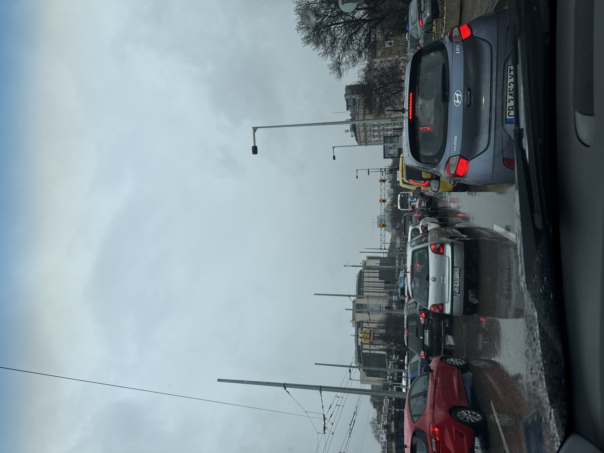 The image depicts a multi-lane urban traffic jam in Sofia, Bulgaria, observed from inside a vehicle. The location is identifiable by the prominent National Palace of Culture (NDK) building in the background, visible on the left with a large "McDonald's" sign on its facade. To the right, another building displays a "ROLEX" sign. The scene is set under an overcast sky, with the asphalt road visibly wet, reflecting vehicle lights and overhead structures, indicating recent or ongoing rain.
Multiple vehicles are stationary or moving slowly in several lanes. In the immediate foreground, the rear of a light blue Hyundai i10 (Bulgarian license plate CB 3452 VT) is visible, its red brake lights illuminated. To its left, a silver Volkswagen Polo (license plate CB 4301 CB) also shows illuminated red brake lights. Further left, a red hatchback is partially visible. Behind the Hyundai, a yellow taxi van is stopped. Overhead lines for trolleybuses and streetlights are present, crisscrossing the sky. Traffic lights in the distance are red. The lower portion of the image shows the interior of the observing vehicle, including the dashboard and the base of the windshield wipers. No pedestrians are visible.