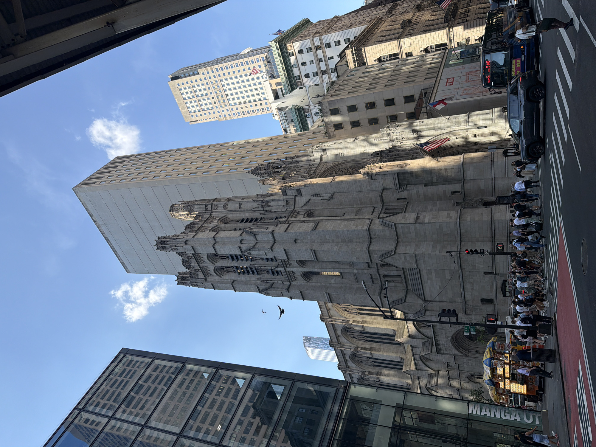 The image shows a street scene in New York City, USA, looking up at the buildings. The focus is on Trinity Church, a large Gothic Revival church. To the left of the church is a modern glass building reflecting other buildings. Above Trinity Church are multiple skyscrapers. A number of people are standing on the sidewalk next to the crosswalk. There are vehicles and a hot dog stand on the right of the image. The sky is blue with few clouds. A single bird is flying in the sky.