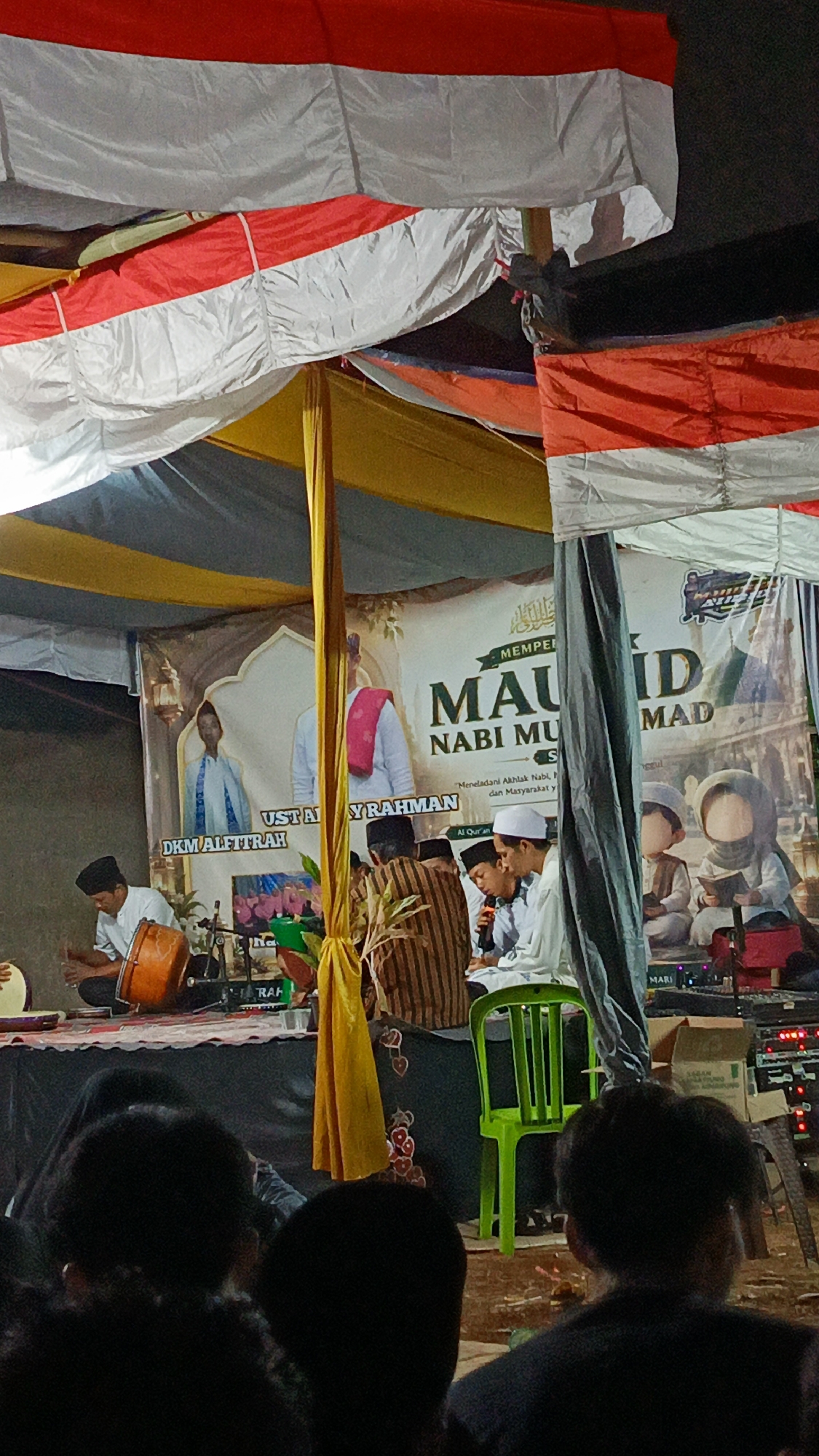 The image captures an outdoor event at night in Margajaya, Indonesia, featuring a religious gathering. The scene is illuminated by artificial lights, casting a warm glow. In the foreground, several individuals are seated, their faces obscured by the dark, as they observe the proceedings. A large banner in the background displays Islamic calligraphy and depicts figures, hinting at the theme of the event. The backdrop is adorned with red and white fabric, possibly representing the Indonesian flag, suggesting a patriotic or nationalistic element to the occasion. On a raised platform, several men are actively engaged. One man is playing a musical instrument, likely a drum, while others are speaking into microphones, possibly delivering a sermon or participating in a religious discussion. The overall atmosphere appears to be one of solemnity and community participation, centered around a significant religious observance.