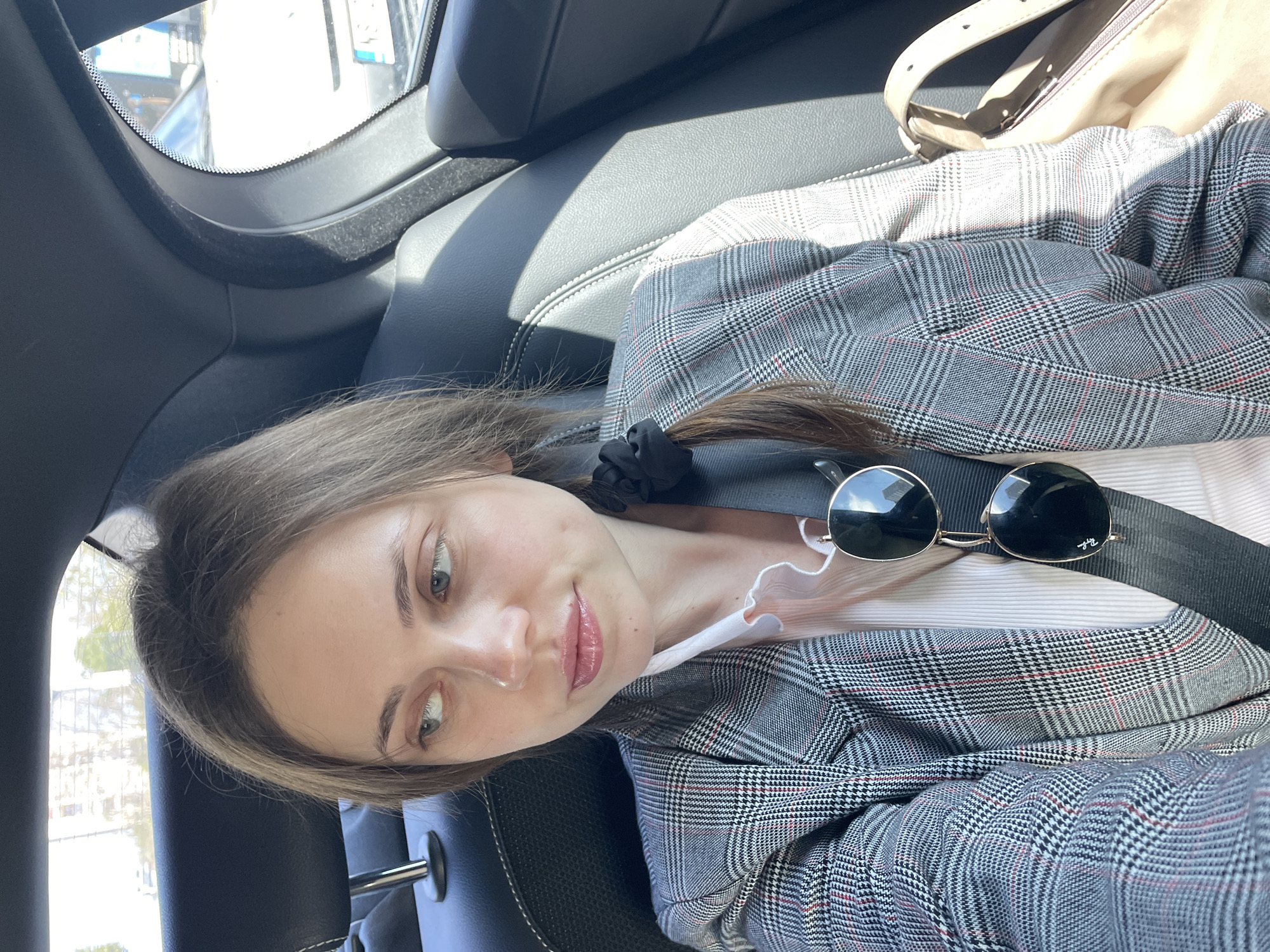 A young woman is seated in the back of a car, wearing a gray plaid blazer, a light pink blouse with a ruffled collar, and a black scrunchie in her hair. A pair of round sunglasses rests on her lap, and a seatbelt is across her chest. A beige purse is visible in the upper right corner of the frame. The car's interior is dark gray leather. Outside the window, blurry shapes suggest a street scene, possibly within the city of Novi Sad, Serbia.