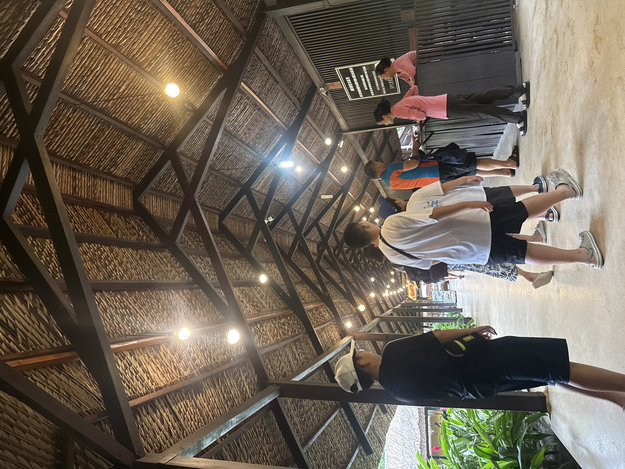 The image depicts an outdoor covered walkway with a thatched roof supported by dark wooden beams. The walkway is lined with lights and appears to be a thoroughfare in a public area. In the foreground, several people are walking along the path. On the left, a young person wearing a black t-shirt, black shorts, and a white baseball cap is walking away from the camera. To their right, a group of people are walking towards the camera. On the far right, two individuals, appearing to be staff, are standing behind a counter or information desk. The environment suggests a tropical or subtropical location, possibly a resort or a tourist attraction. The signage visible on the counter and on the wall behind it is in Vietnamese, indicating the location is in Vietnam. Considering the typical architecture and the presence of a counter for ticketing or information, this could be an entrance or an information point at a place like VinWonders Nha Trang or a similar tourist destination in Nha Trang, Vietnam.