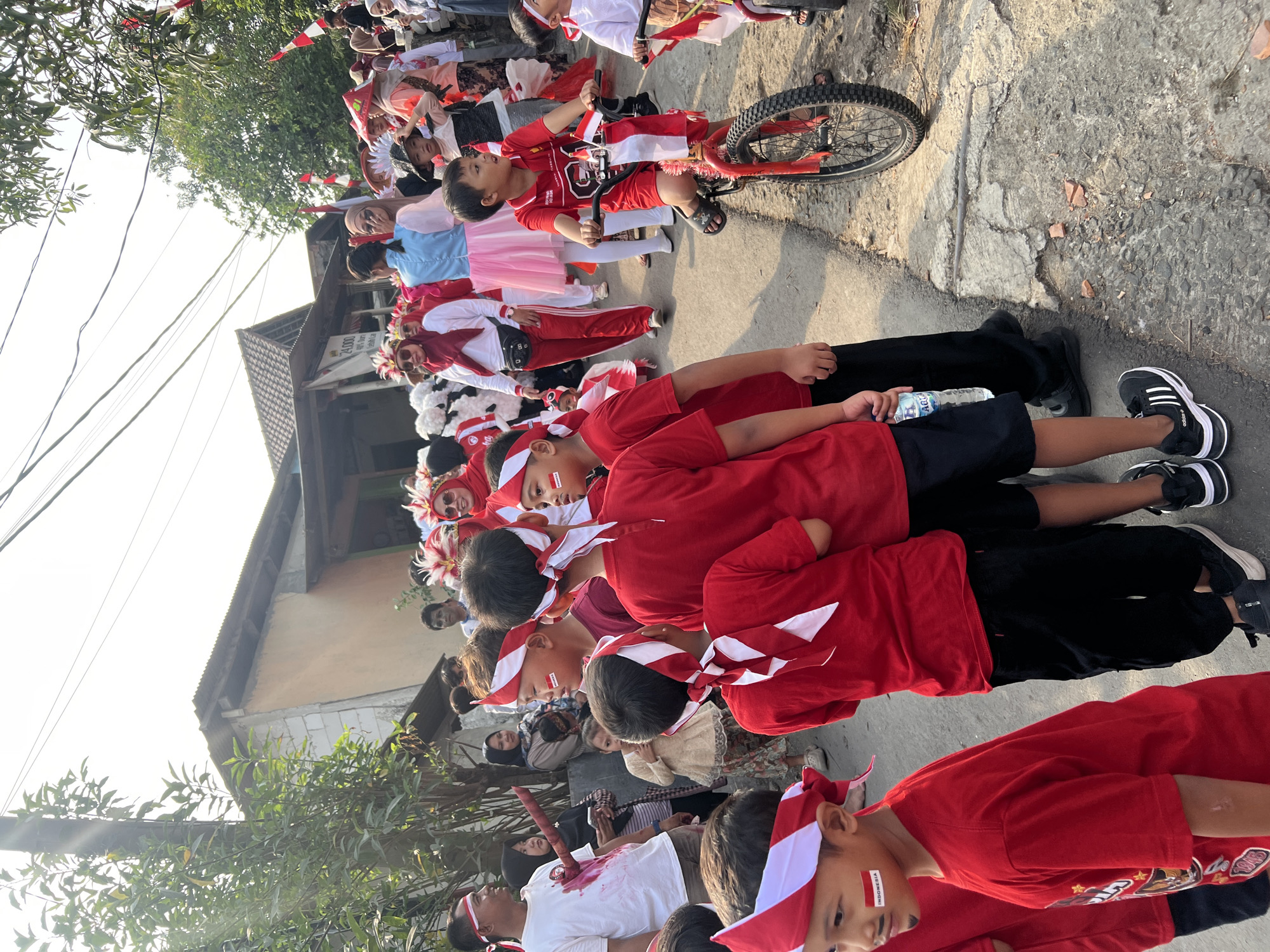 In Klapanunggal, Indonesia, a procession of children and adults, many adorned with red and white headbands and face paint, walk along a paved street under a bright sky. Several children are dressed in red t-shirts and black shorts, while some adults wear red and white attire, and others in festive costumes with feathered headdresses. A young boy rides a red bicycle decorated with Indonesian flags. The scene appears to be a celebration or parade, possibly for a national holiday, with spectators lining the street. Buildings with corrugated metal roofs and muted-colored walls are visible in the background. The overall atmosphere is lively and festive.