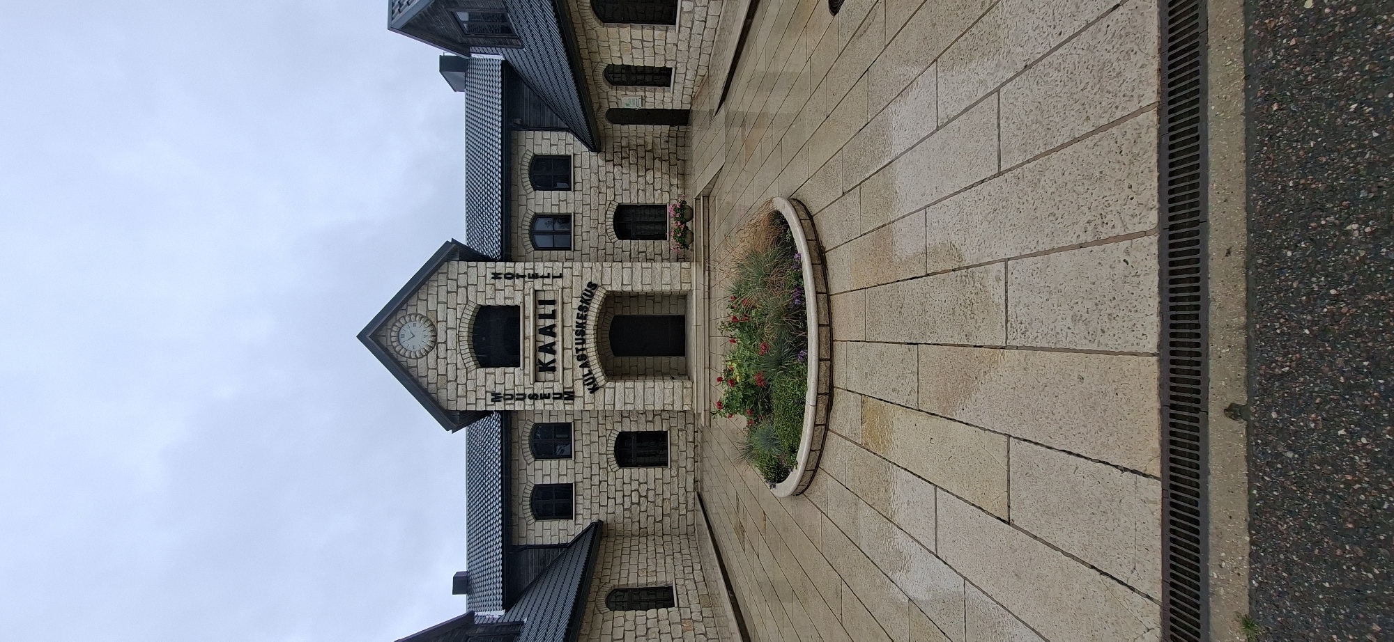 A front-facing, eye-level, medium shot captures the Kaali Museum and Visitor Centre in Kaali, Estonia. The building, constructed of light-colored stone blocks, features a prominent central section with a dark-tiled roof and a clock face above an arched entrance. The words "MUSEUM," "KAALI," and "HOTEL KULASTUSKESKUS" are displayed in black lettering on the facade. To the left and right of the central structure are lower, symmetrical wings also made of stone, with dark-tiled roofs and black-framed windows. A circular flower bed with red and pink flowers and ornamental grasses is situated in the foreground, surrounded by a paved courtyard of large, rectangular stone tiles. The sky is overcast and the pavement appears wet, suggesting recent rain. A dark metal grate runs along the bottom edge of the paved area. The scene is devoid of people.