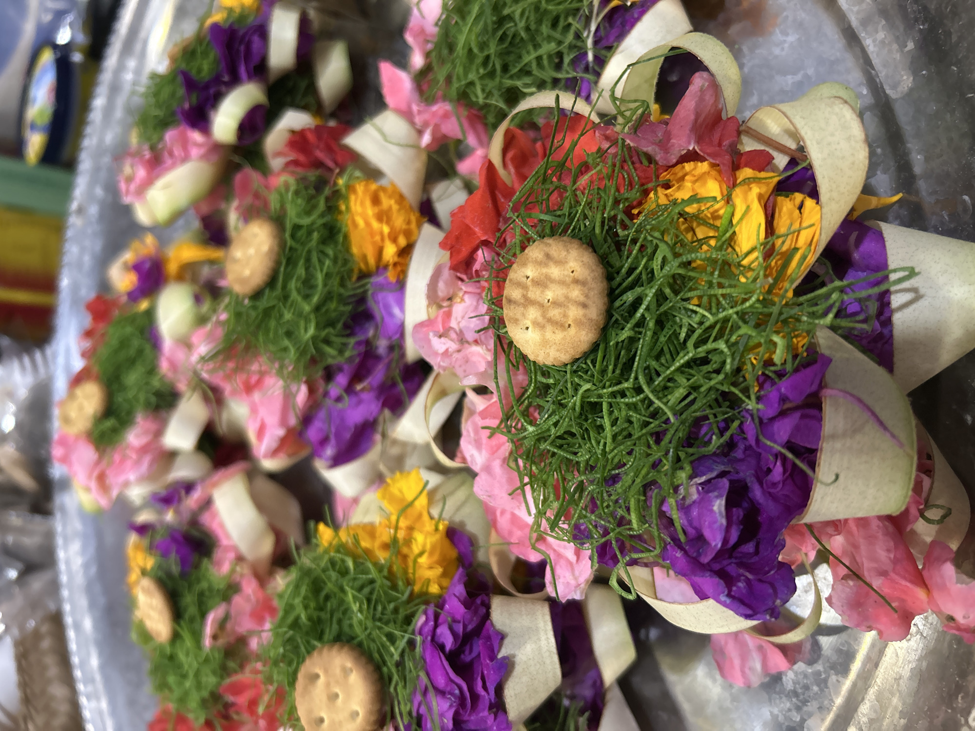 The image displays a close-up of several small, intricately arranged offerings, likely placed on a metal tray. Each offering appears to be a small cup or basket fashioned from a light-colored material, possibly woven leaves or palm fronds. These containers are filled with a vibrant mix of colorful flower petals, including shades of pink, purple, yellow, and red. Nestled on top of the greenery and flowers in some of the offerings is a small, round cracker with visible holes. Thin, light-colored ribbons are intertwined with the floral arrangements. The background is blurred, suggesting a shallow depth of field, and hints at a metallic surface beneath the offerings, consistent with a tray. The overall impression is of a cultural or religious offering, potentially from a market or ceremony. Given the location information, this scene could be from the bustling markets of Sukawati, Indonesia, known for its traditional crafts and offerings.