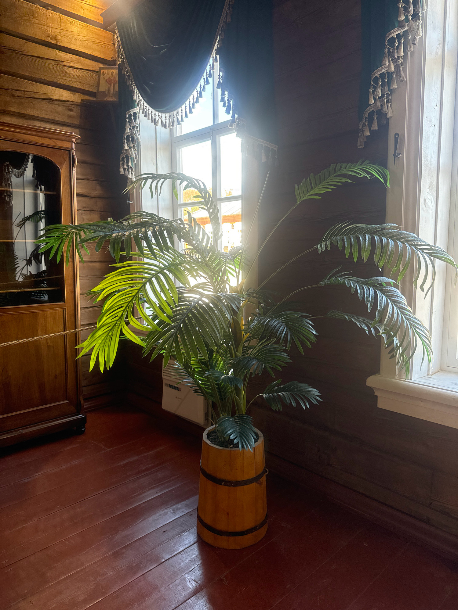 The image depicts an interior scene in the Museum of Wooden Architecture in Taltsy, Russia. A large artificial palm plant in a wooden barrel planter stands in the center of the frame. The plant's fronds, some illuminated by sunlight streaming through a window, are lush and green. To the left of the plant is a tall, wooden cabinet with a glass door. The walls are made of dark, rough-hewn logs. Heavy, dark velvet curtains with ornate tassels hang near the window, which is large and divided into panes. The floor is made of dark red wooden planks. A white radiator is visible behind the plant. An Orthodox icon is displayed on a small shelf on the wall above the cabinet. The overall impression is of a rustic, yet furnished interior, likely part of a historical exhibit.