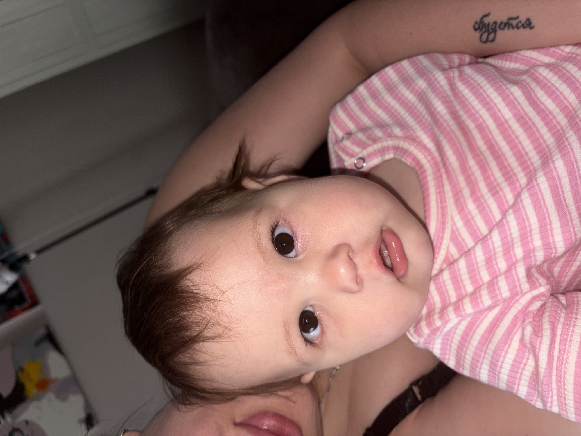 A close-up shot depicts a baby with brown hair and large brown eyes, lying on someone, wearing a pink and white striped shirt. The person holding the baby is partially visible, with a tattoo on their arm in Russian, and some underclothing showing. The background is blurred, showing a wall with a dresser and some items on top of the dresser. The location and city are unknown.