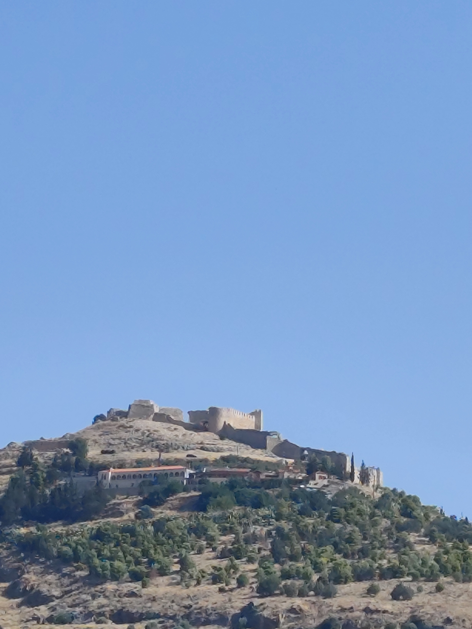 The image depicts the Larissa Castle located in Argos, Greece, atop a hill against a clear blue sky. The castle walls, constructed of stone, are visible on the summit. Below the castle, a long building with an arched facade and a terracotta roof is situated on the hillside, surrounded by lush green trees and vegetation. The terrain is arid and rocky. There are no people or discernible actions in the image.