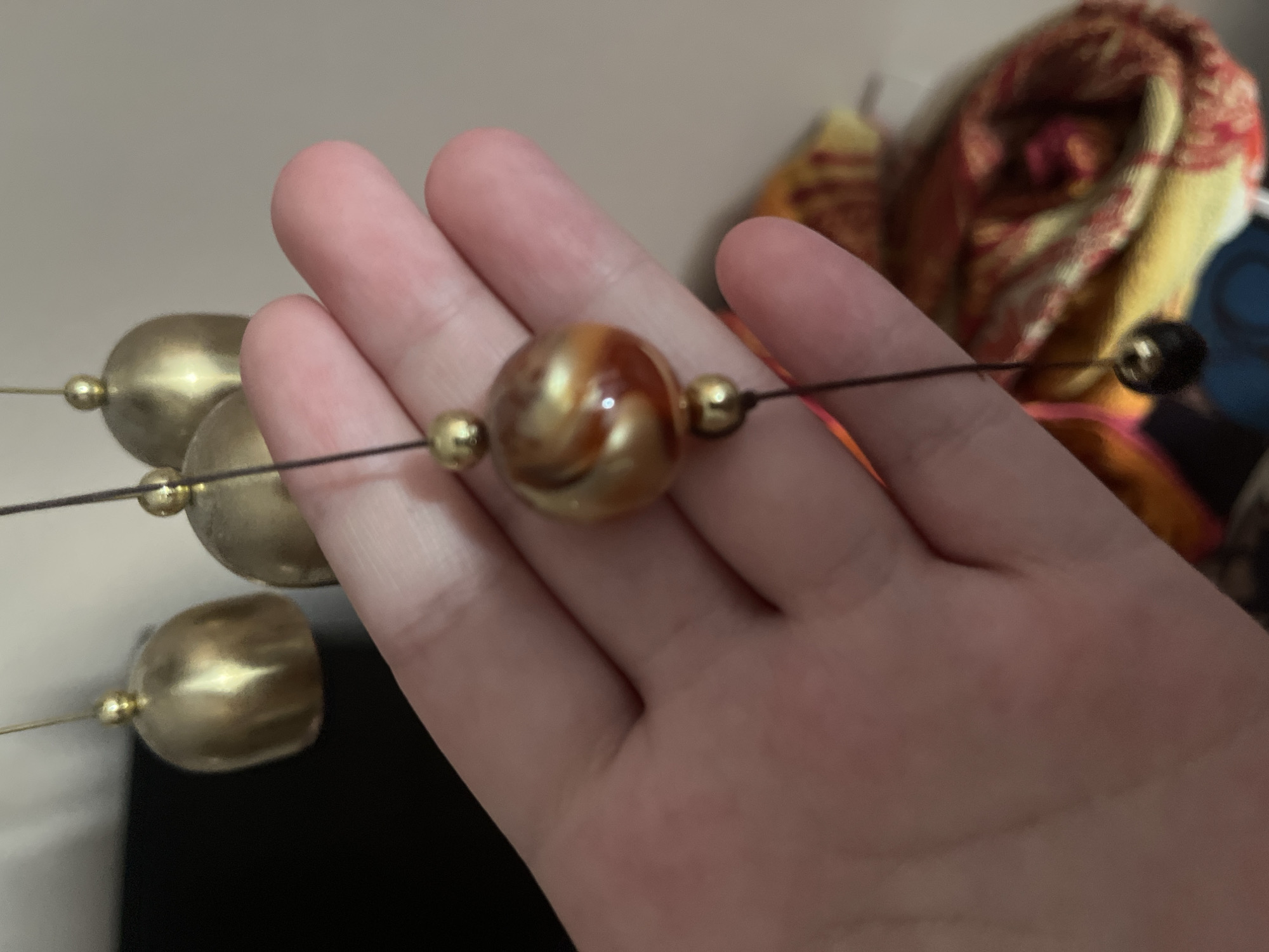 In an indoor setting, possibly a home, a person's hand is visible, gently holding a string of decorative beads and bells. The beads are a mix of gold, with one prominent bead featuring swirls of gold and amber. Several small, golden bells, reminiscent of wind chimes, hang above the beads, suspended by thin wires. The background is blurred, but suggests a softly lit room with some fabric items, perhaps a scarf or tapestry, in warm tones of yellow, red, and orange. The overall impression is one of delicate ornamentation and a moment of quiet appreciation.