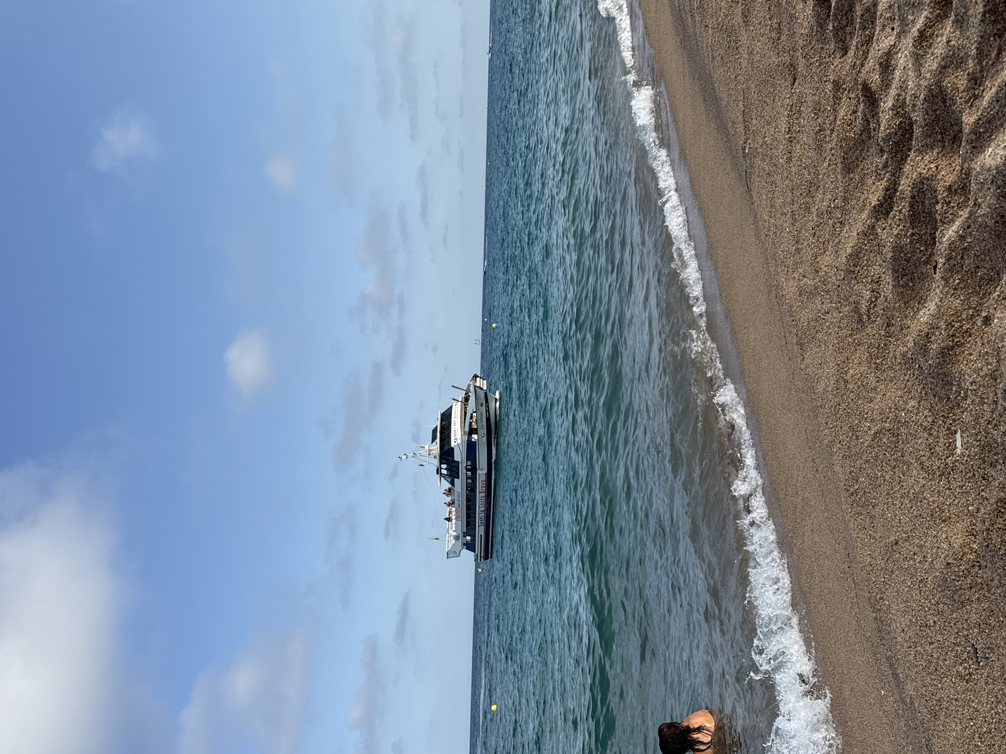 The image depicts a sunny day at a beach in Blanes, Spain, with a boat sailing in the distance. The foreground shows a sandy beach with gentle waves lapping at the shore. A person is partially visible in the water on the left side of the image. In the middle ground, a white and blue boat, labeled "SANT ILLA COSTA BRAVA," is sailing on the turquoise water, with several passengers visible on its deck. The background features a clear blue sky with scattered white clouds. The overall scene conveys a peaceful and recreational atmosphere, typical of a coastal summer day.