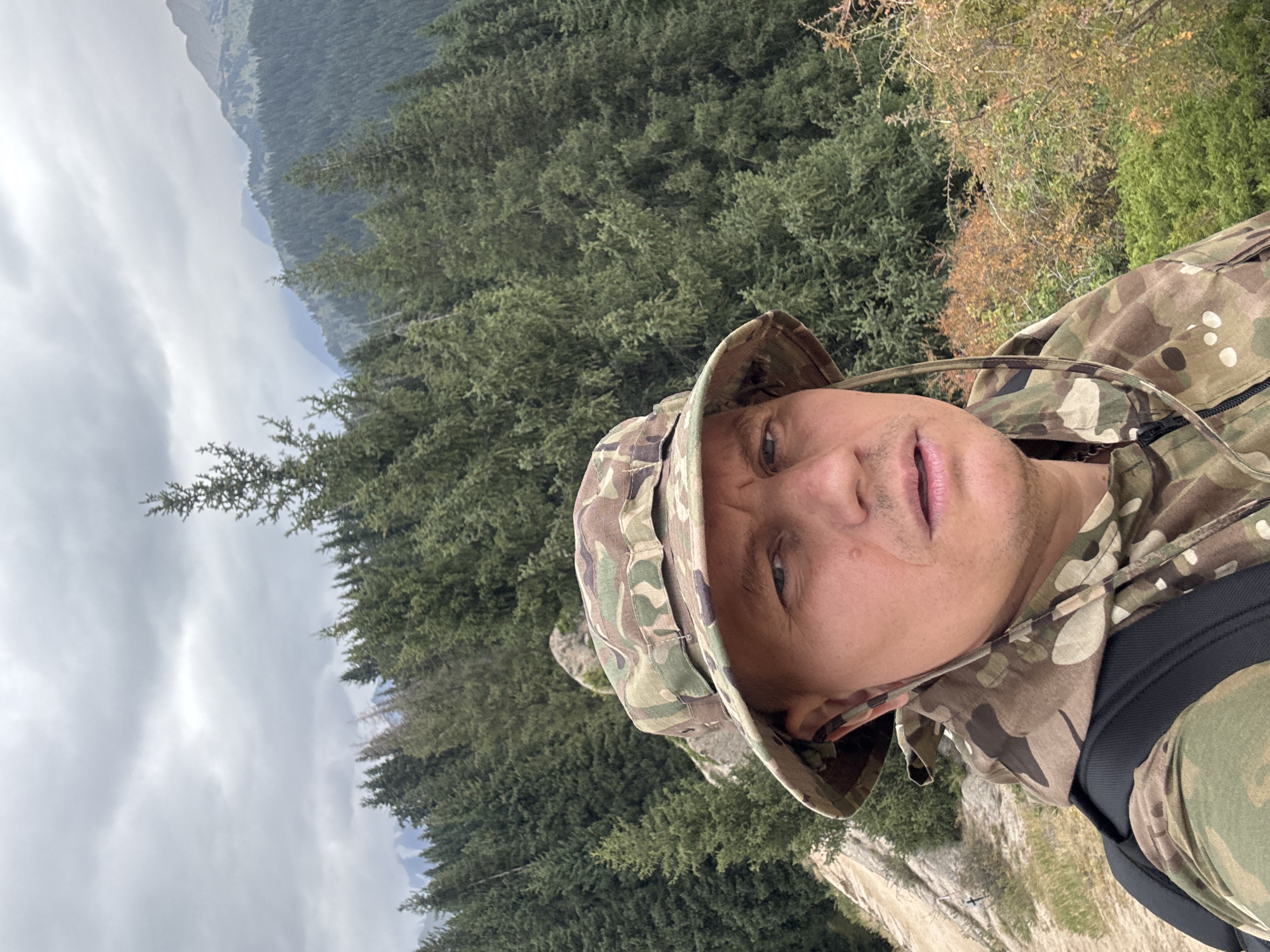 A man wearing a camouflage bucket hat and jacket takes a selfie on a mountain ridge overlooking a dense forest of evergreen trees. The man's face is partially visible, and he appears to be looking directly at the camera. In the background, the forest extends down the mountainside, and further in the distance, several layers of hazy mountains are visible under a cloudy sky. The man has a backpack on. The terrain directly to his left is a rocky, uneven slope. This location is in the mountains above Almaty, Kazakhstan.
