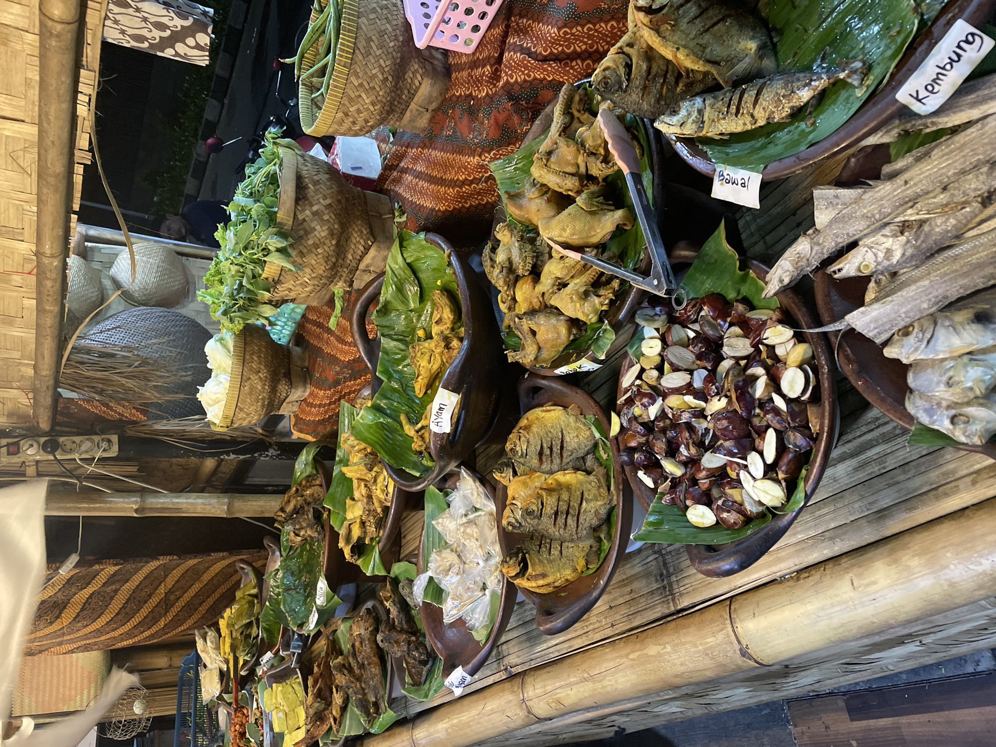 The image shows a food stall at night in North Jakarta, Indonesia. Several bowls of food are displayed on a bamboo table, including fried fish labeled "Bawal" and "Kembung", fried chicken labeled "Ayam", and other foods. A basket with vegetables is on the left. A sign with the word "Nila" is visible in one of the bowls. Some bowls are lined with banana leaves. Tongs are visible in one of the food bowls.