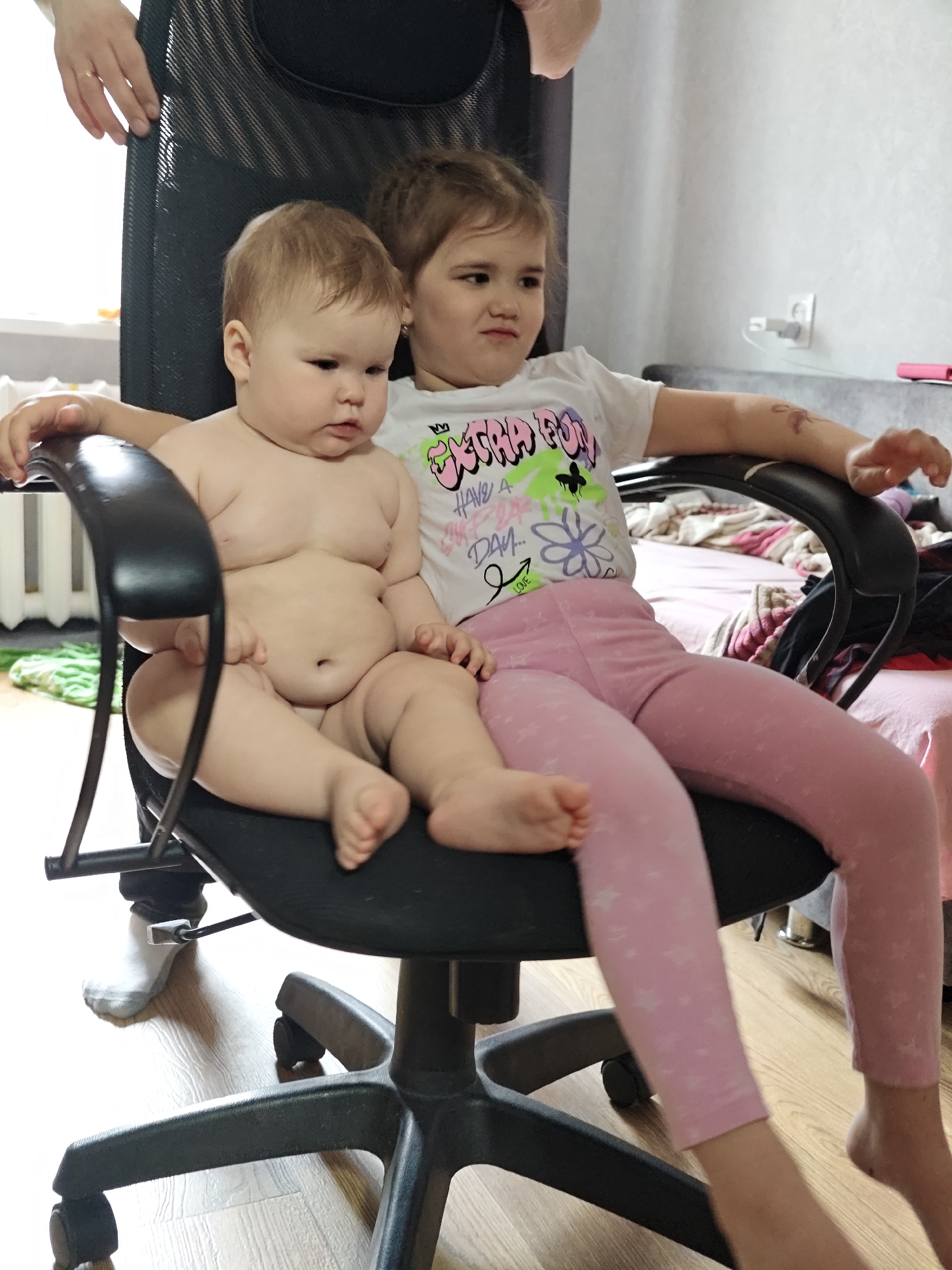 Two young children sit in a black office chair. The younger child, a baby, is unclothed from the waist up and sits in front. The older child wears a white t-shirt with a colorful graphic design and pink leggings with white stars and sits behind, with her arms resting on the chair's arms. A person wearing black pants and a light-colored sock stands near the chair. The chair is positioned on a light wood floor. In the background, there is a bed with pink and white striped sheets and a white wall with an electrical outlet. The location is most likely Chaikovsky, Russia, based on the prompt.