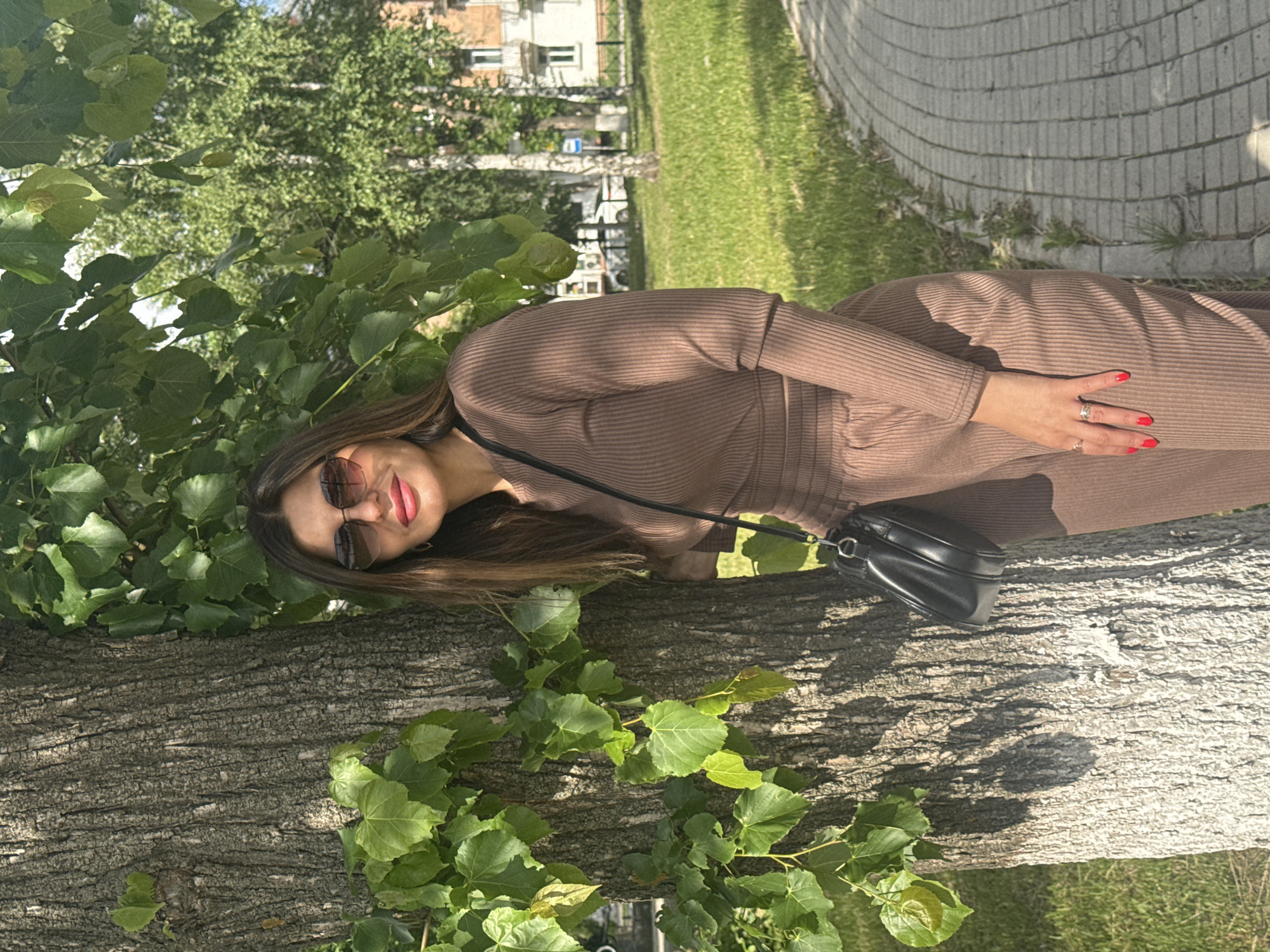 A woman is posing in front of a tree. She is wearing a brown dress and sunglasses, and she is carrying a black purse. The location is a park in the city of Votkinsk, Russia, which is evidenced by the typical architecture and landscaping of the Russian provinces. The setting is outdoors during the day. The background includes grass, trees, and buildings. The woman is interacting with the environment by leaning on the tree.