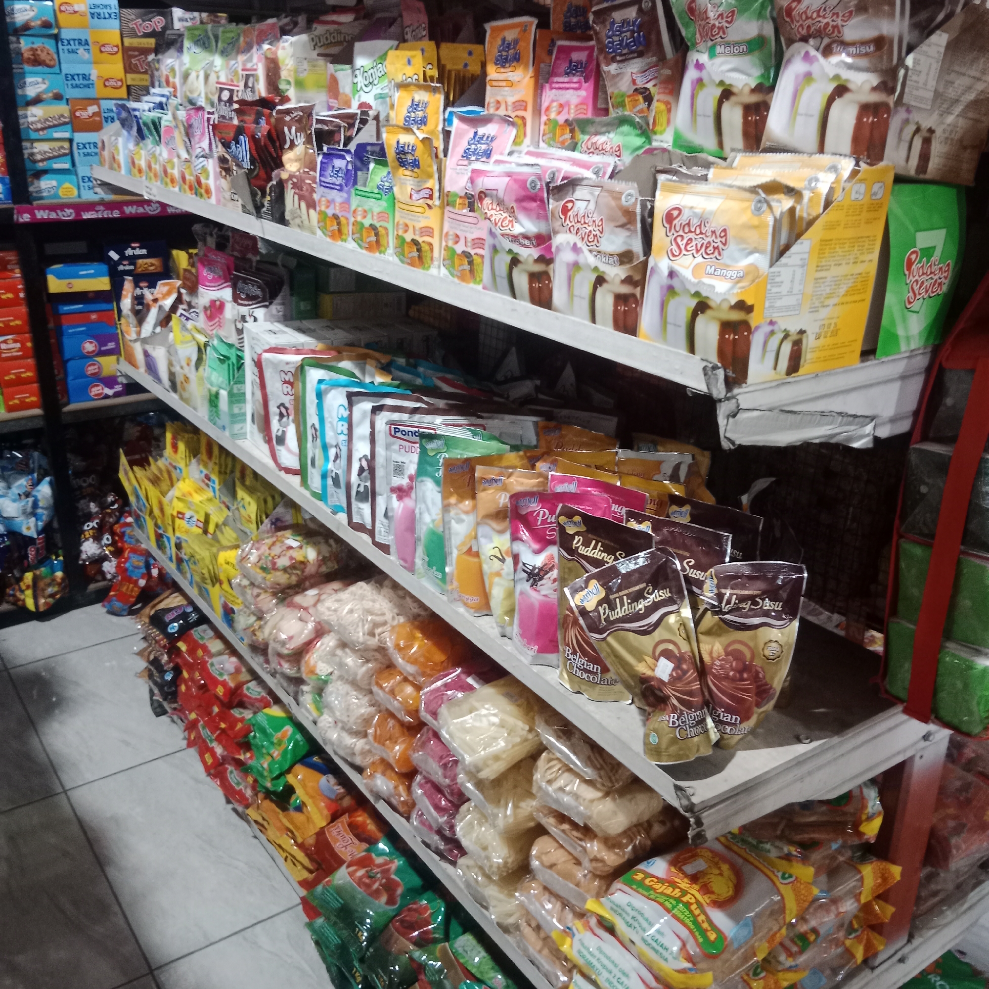 The image shows a retail display of packaged food products, primarily in a convenience store or minimarket. The products are arranged on multiple shelves. On the upper shelves, there are various pouches of pudding mixes, some labeled "Pudding Seven" with flavors like Mango, Tiramisu, and Melon. There are also packages of what appear to be cookies or biscuits. Lower shelves display more packaged food items, including smaller pouches of pudding, snacks like crackers or crisps in plastic bags, and boxed items. Some products have visible text in Indonesian. The overall environment appears to be indoors, with white tiled flooring visible in the lower portion of the image, indicating a commercial setting. No people are visible in the frame. The lighting suggests an interior store environment. The products are diverse, with a focus on desserts and snacks. The store appears to be located in Tegowanu Wetan, Indonesia, as indicated by the context provided.