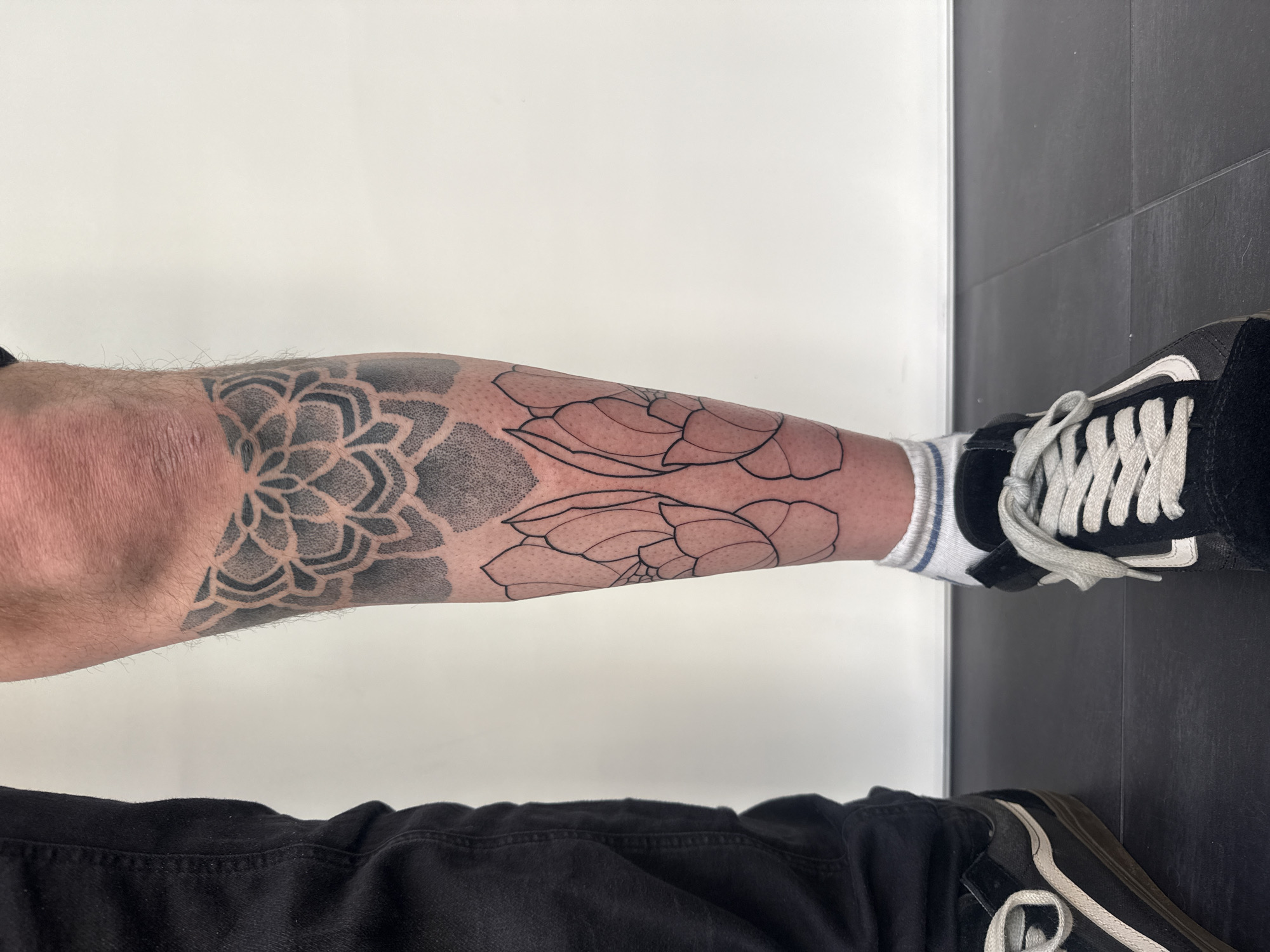A person's lower left leg is visible from the knee down, against a white wall and a dark tiled floor. The leg has a tattoo that starts with a patterned, mandala-like design near the knee, transitioning into two large, outlined floral shapes lower down. The person is wearing black pants and black Vans sneakers with white laces. A white sock with blue stripes is visible above the right sneaker. The environment appears to be an indoor studio or room.