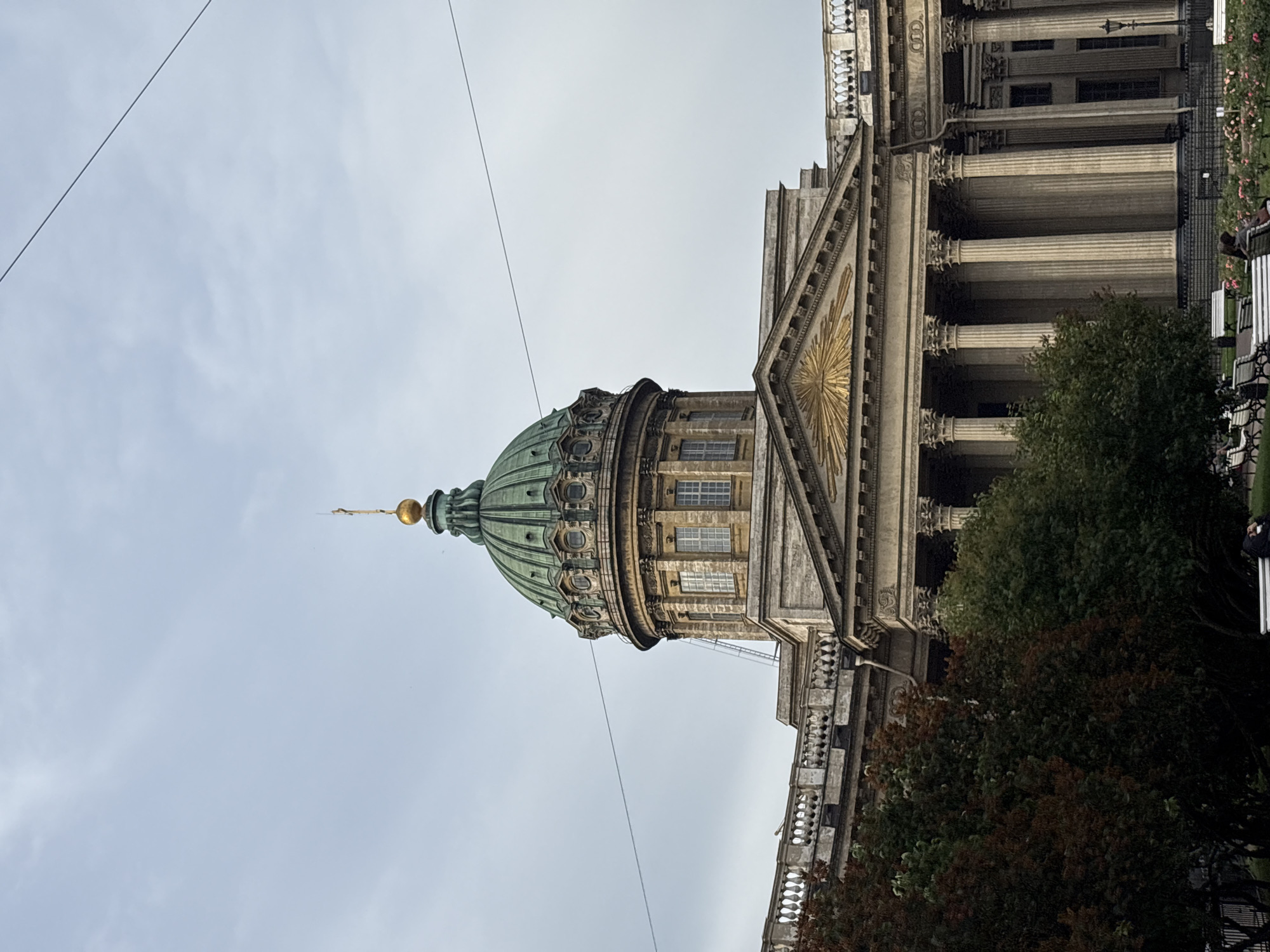 The image shows the Kazan Cathedral, a prominent landmark in Saint Petersburg, Russia. The cathedral features a large, green dome topped with a golden spire and cross. The building's facade is adorned with classical columns and ornate architectural details. In the foreground, lush green trees with hints of autumnal color are visible, partially obscuring a park area with benches where several people are seated. The sky is overcast with a light grey hue. The image captures a serene, somewhat muted atmosphere, typical of a cloudy day in Saint Petersburg.