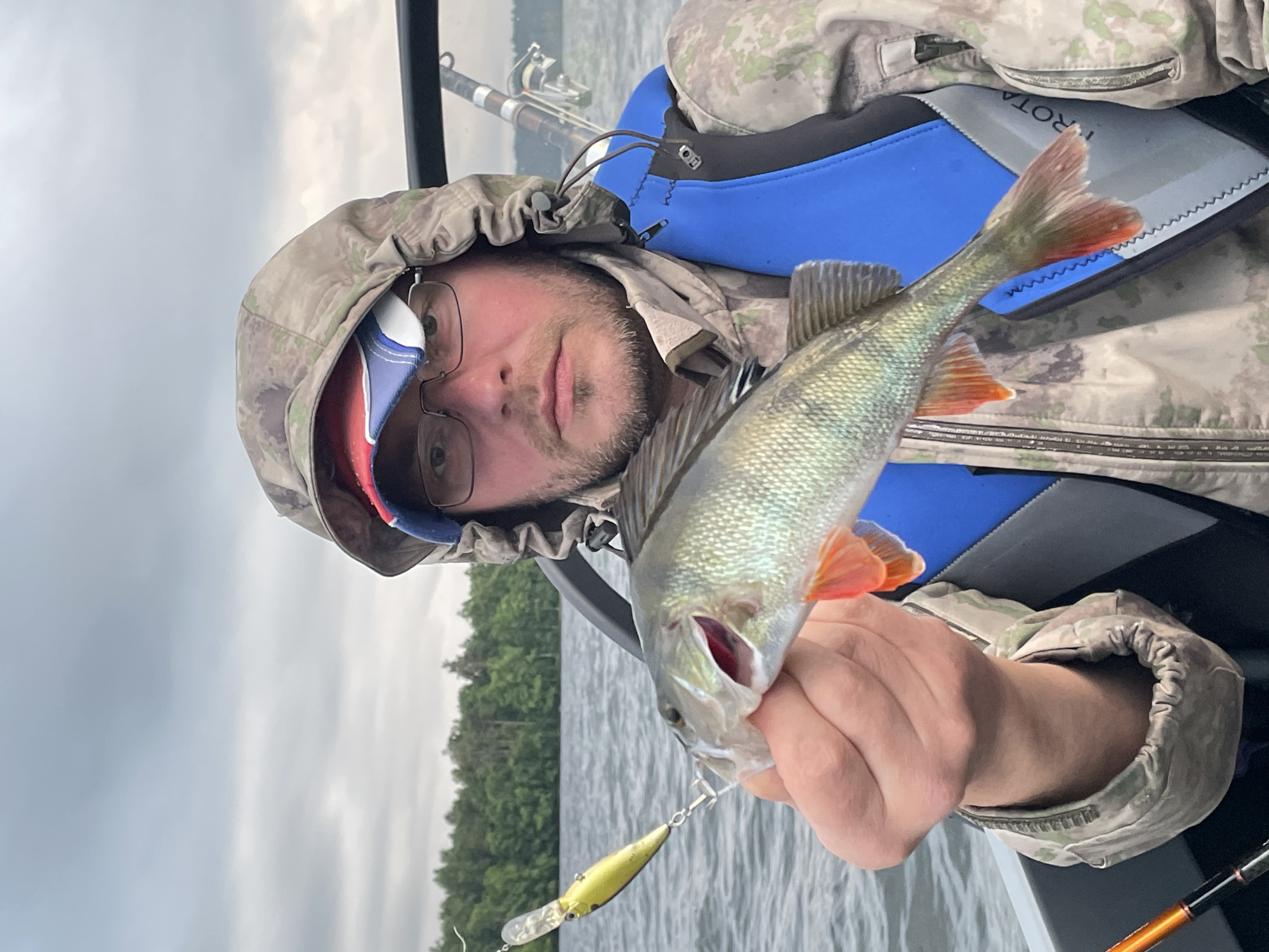 A man in a camouflage jacket and blue life vest is holding a freshly caught fish. The fish, a perch, has its mouth open and is dangling from a fishing lure. The man is wearing glasses and a hat. He is in a boat on a lake with trees on the horizon. The sky is cloudy. The scene is likely in Yablonovka, Russia.