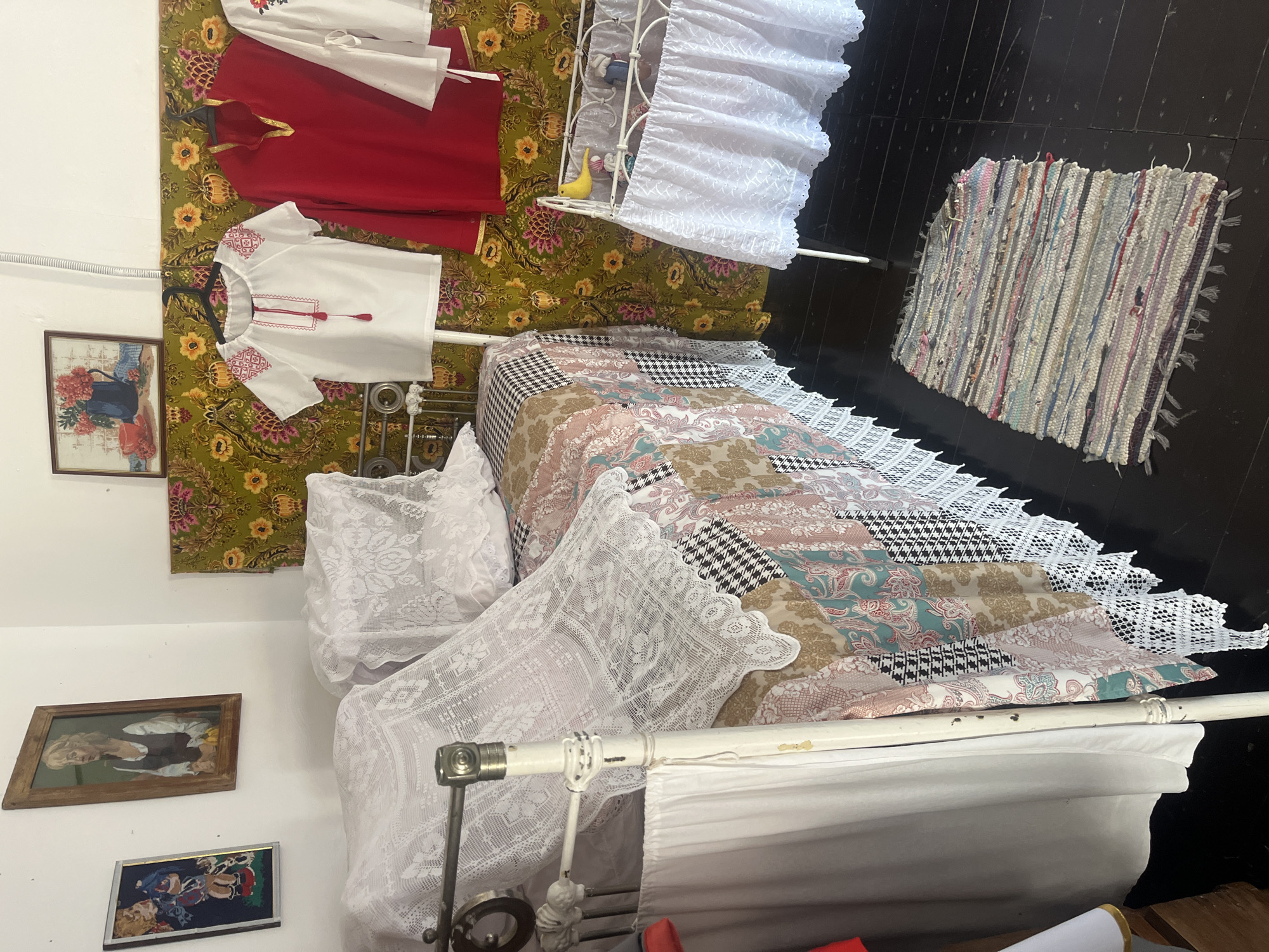 The image shows a bedroom in the Taman State Museum, Russia. The room features a white metal-framed bed with a patchwork quilt and lace coverings. Several framed pictures hang on the wall, including a portrait of a woman and a still life of flowers in a blue vase. Colorful fabric with floral patterns covers a portion of the wall behind the bed. Two traditional embroidered shirts and a red garment are hanging on hangers. A small white crib with lace trim stands near the bed, adorned with a yellow bird figurine and two small dolls. A woven rug lies on the dark wooden floor.