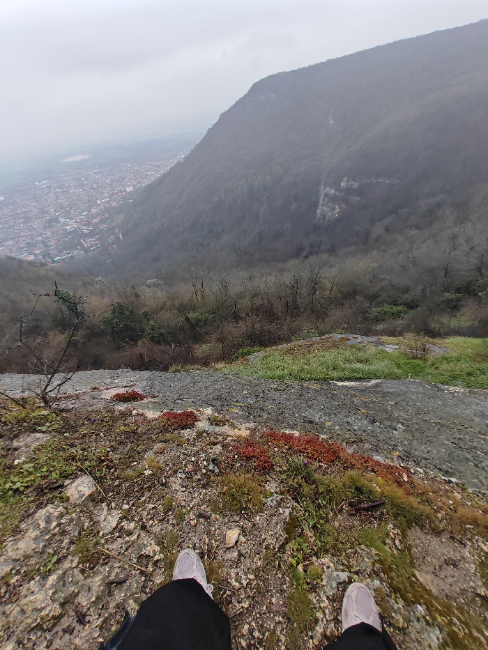 The image depicts a first-person view from a high vantage point overlooking the city of Shumen, Bulgaria, under an overcast sky. In the immediate foreground, a person's feet, clad in light grey sneakers and dark trousers, are visible standing on a rocky, uneven surface covered with patches of green moss and distinctive clusters of reddish-brown vegetation. This surface appears to be the edge of a steep incline or cliff. Below and extending into the midground, a densely forested hillside, primarily composed of bare deciduous trees, slopes sharply downwards into a valley. Visible through the trees are areas of exposed rock faces. In the background, the city of Shumen is sprawled across the valley floor, characterized by numerous buildings with reddish roofs. To the right, a large forested mountain, part of the Shumen Plateau, rises prominently. The distant horizon is hazy under a uniformly grey, overcast sky. No other individuals or specific actions are discernible beyond the implicit act of standing and observing the vista.