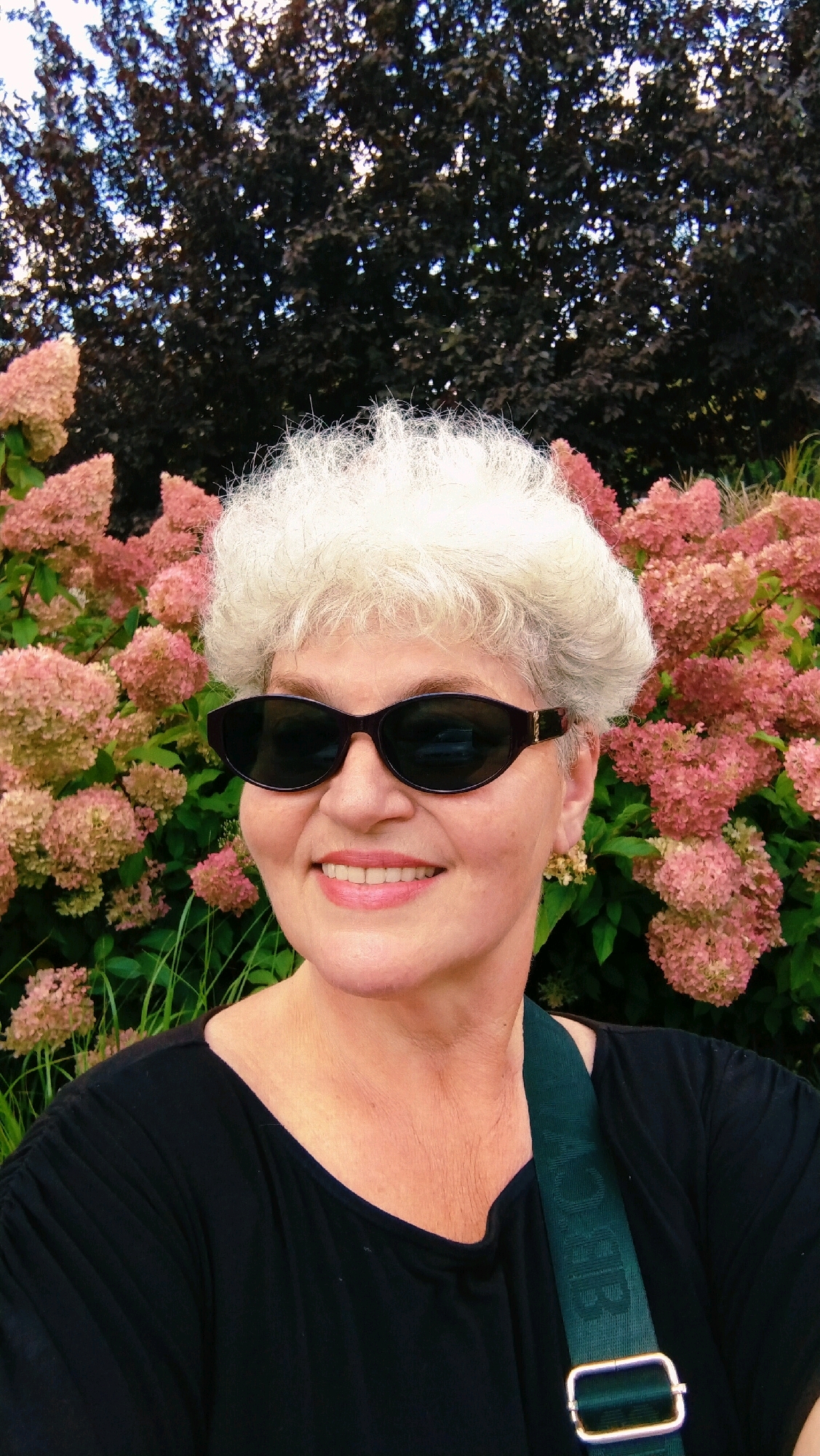 A woman with short, white, curly hair and fair skin, wearing sunglasses and a black shirt, stands in front of a bush with pink flowers. She is smiling. A dark green strap with embossed lettering is visible across her chest. The background consists of dark foliage above and the pink flowers and green foliage below. The scene is outdoors, likely in a park or garden. The image does not provide enough information to determine if it is located in Horishni Plavni, Ukraine.