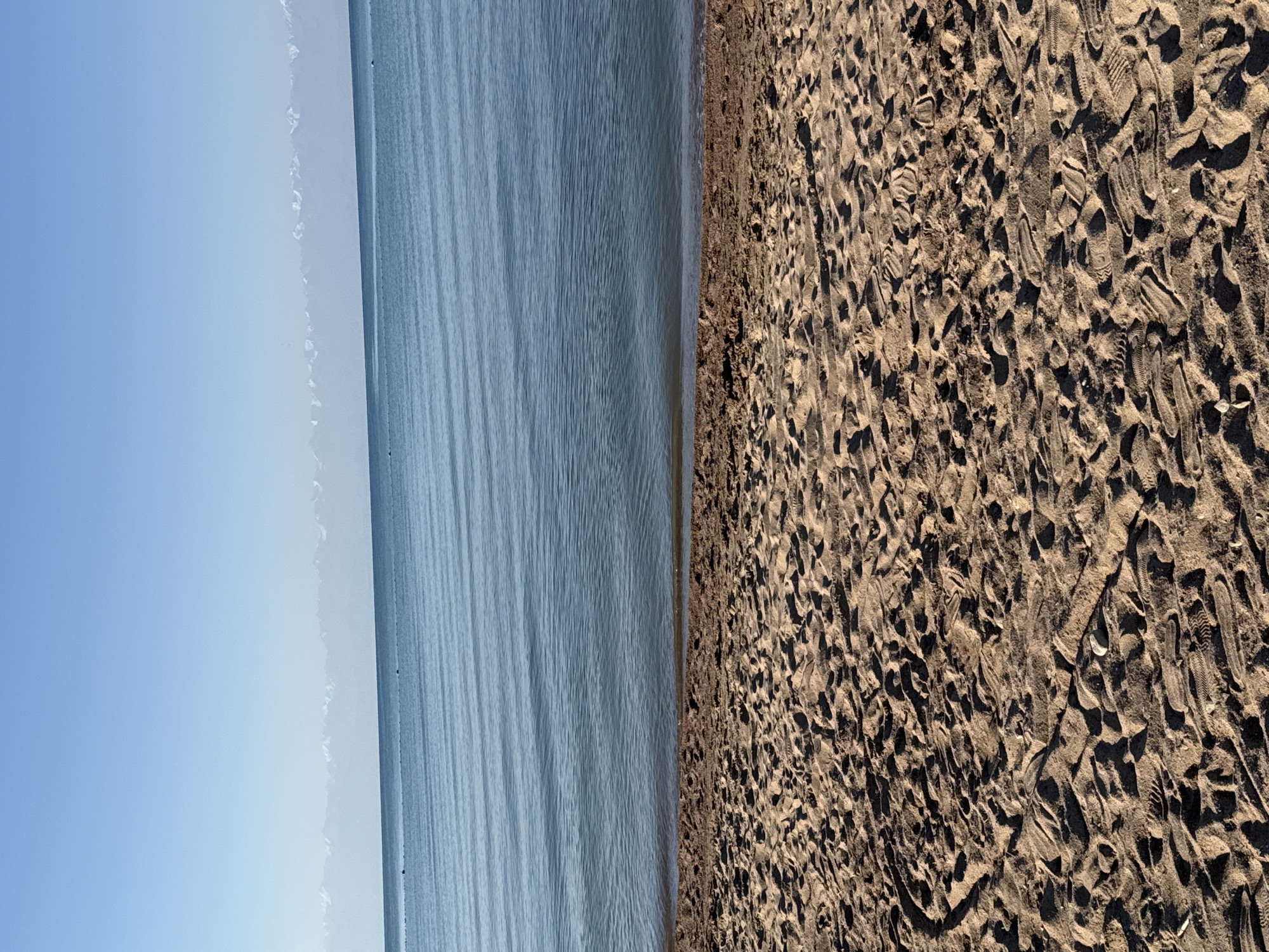 The image depicts a serene natural landscape featuring a beach and a body of water, likely Issyk-Kul Lake, given the geographical context of Sary-Oy, Kyrgyzstan. In the foreground, a sandy beach is visible, marked by numerous footprints and some debris, suggesting recent human activity. The sand itself appears to be a light brown color. Beyond the beach, the calm blue water of the lake stretches to the horizon, reflecting the clear sky above. The water's surface is rippled, indicating a gentle breeze. In the distance, a range of snow-capped mountains forms a majestic backdrop against the pale blue sky. The lighting suggests it is daytime, likely morning or afternoon, with the sun casting shadows on the sand. The scene is devoid of people or any structures, emphasizing the natural beauty and tranquility of the location.