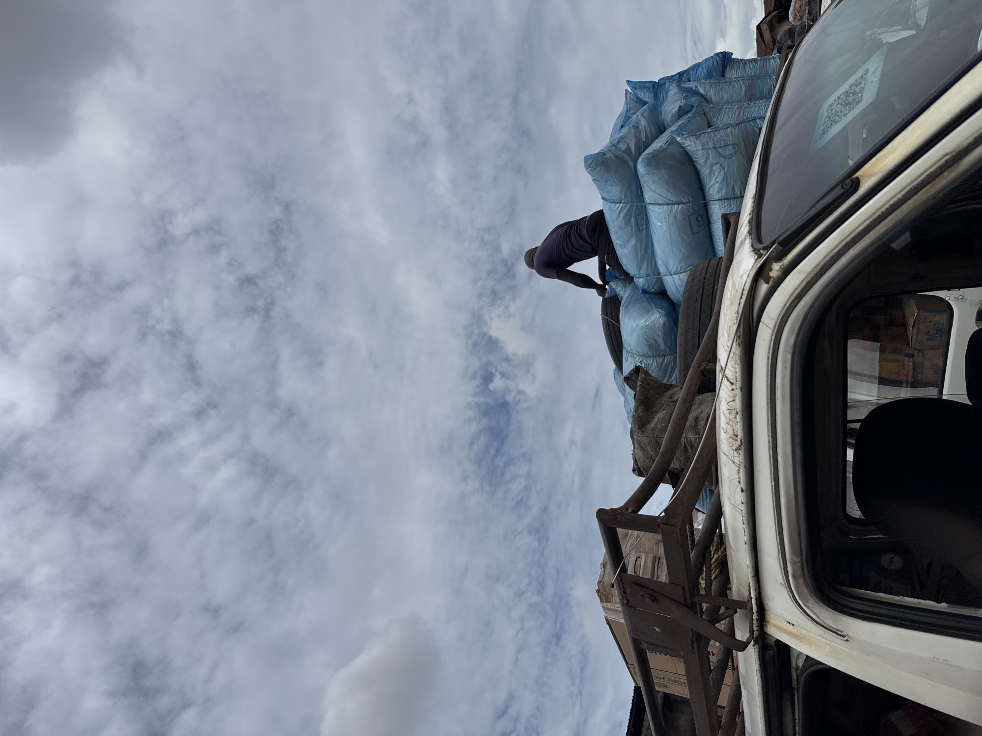 A man is standing on top of a vehicle loaded with blue sacks, possibly loading or adjusting them. The vehicle is white and appears to be heavily laden. It is unclear whether this photo was taken in Onitsha, Nigeria, but the presence of a heavily loaded vehicle is consistent with the transportation of goods often seen in urban areas in Nigeria. The sky is cloudy.