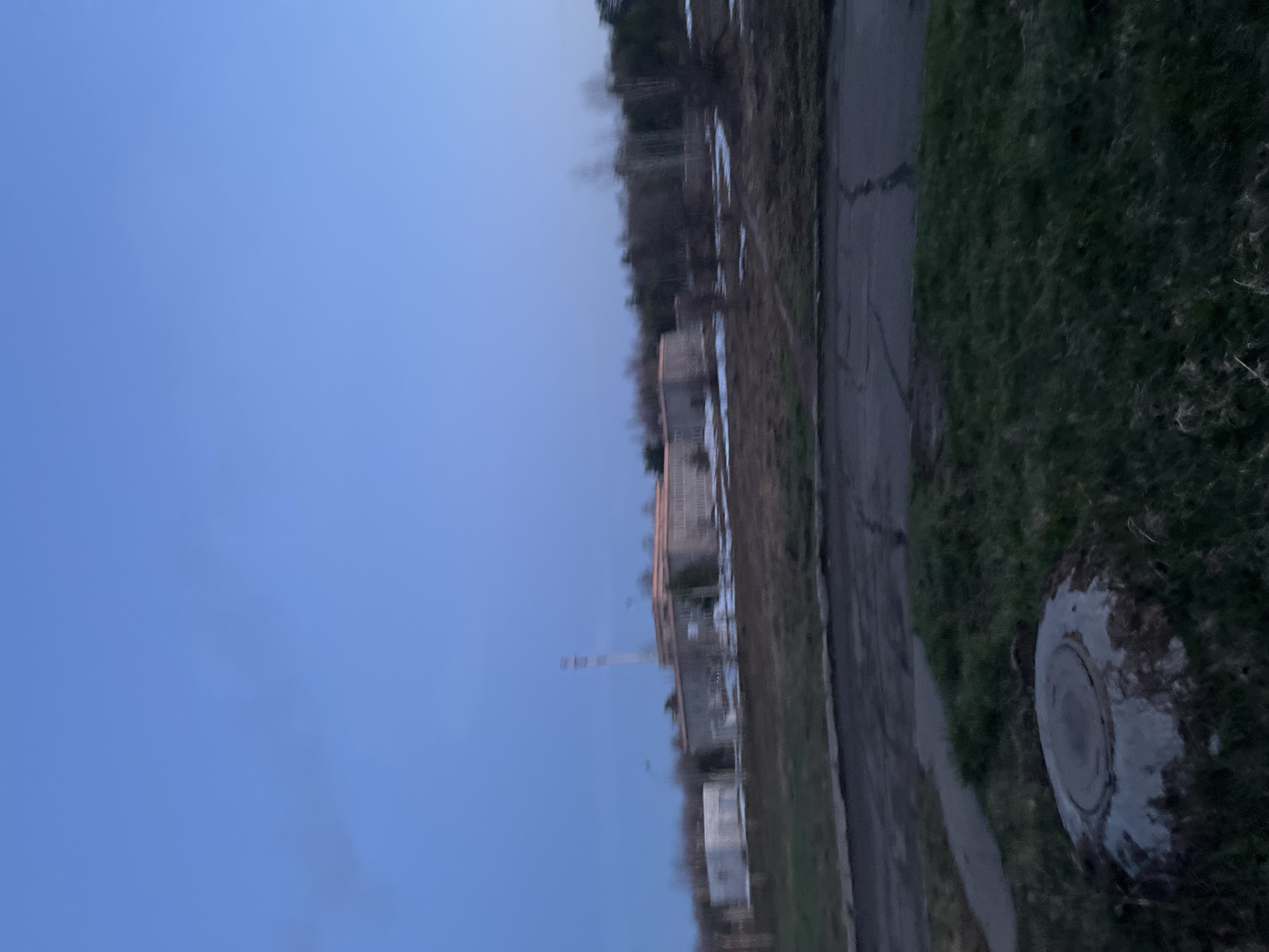 The image depicts an outdoor scene in what appears to be the town of Марковский, Russia. The sky is a clear blue, indicating either early morning or late evening. A grey concrete structure stands parallel to the street, with trees and patches of snow visible in the background. A small road runs along the edge of a grassy area. On the right side of the image, a manhole cover is visible in the grass. There are no people visible in the image.