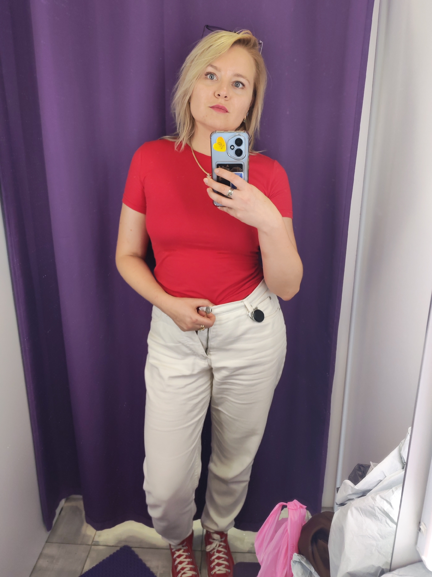 In what appears to be a changing room in a retail establishment in Izhevsk, Russia, a woman with blonde hair stands facing a mirror, taking a selfie with her smartphone. She is wearing a bright red t-shirt and off-white pants, accessorized with rings on her fingers and a necklace. The background consists of a deep purple curtain and a white wall, typical of a fitting room environment. On the floor, a small purple mat is visible. The woman is in the process of adjusting the button or zipper of her pants. The lighting suggests an indoor setting, likely a store.