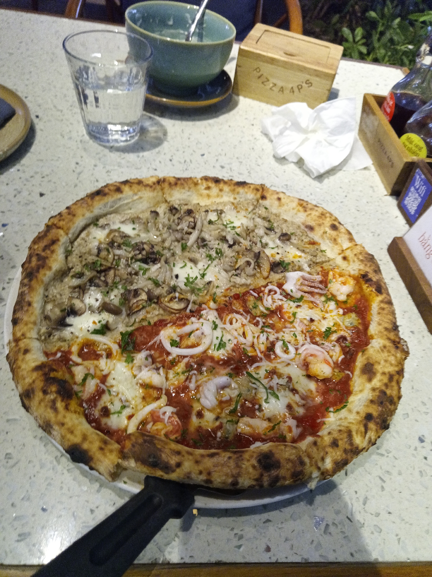 This media file presents a close-up view of a large, freshly baked pizza placed on a white plate at a dining table. No people are visible in the image.
The pizza is divided into two distinct halves with different toppings. The left half features a generous topping of various mushrooms, including darker, smaller varieties and lighter, larger ones, set on a white sauce base, possibly cream or cheese, and garnished with fresh green herbs like parsley. The right half displays a vibrant red tomato sauce, melted cheese, and an assortment of seafood, specifically white calamari rings and pink shrimp, also adorned with green herbs. The pizza's crust is thick, airy, and shows characteristic charring, indicative of a wood-fired oven.
The dining table, with a light-colored, speckled surface resembling granite or quartz, holds several other items. To the upper left, a clear glass of water sits next to a teal-green bowl containing a spoon, appearing mostly empty. In the upper right, a small, light-colored wooden box is clearly labeled "PIZZA 4P'S." Beside it lies a crumpled white napkin. Further right, a wooden caddy holds two dark bottles, likely condiments, with a small yellow circular label attached and a blue sign displaying a QR code and "Wifi." The text "PIZZA 4P'S" is also partially visible on the side of this caddy. A black pizza cutter handle is visible in the lower foreground.
The setting appears to be a restaurant, specifically a branch of Pizza 4P's, a well-known establishment in Vietnam, which aligns with the provided location context of Nha Trang. The background shows hints of green foliage and wooden structures, suggesting an indoor-outdoor or garden-like dining environment. The lighting suggests an evening or indoor dining experience, creating a warm atmosphere. The overall scene depicts a pizza prepared for enjoyment.