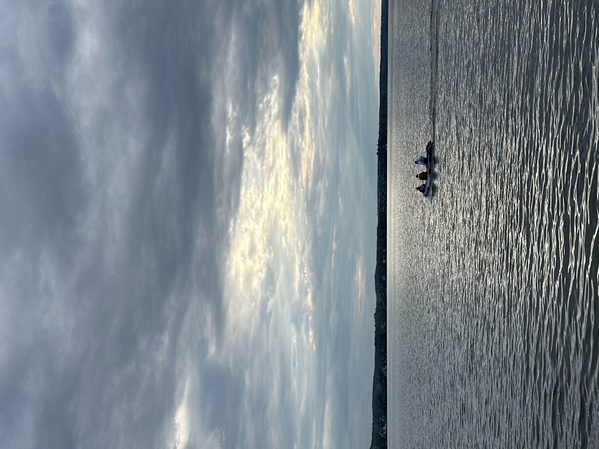 An overcast sky looms over a calm body of water. Sunlight breaks through the clouds in streaks, illuminating the surface of the water with a shimmering reflection. In the distance, a dark, wooded shoreline is visible, dotted with hints of structures, suggesting a settlement. A small inflatable boat with three individuals aboard motors across the water, leaving a slight wake. The overall mood is serene, with the dramatic sky contrasting with the peaceful water and the distant, muted landscape. The location is the city of Votkinsk, Russia.