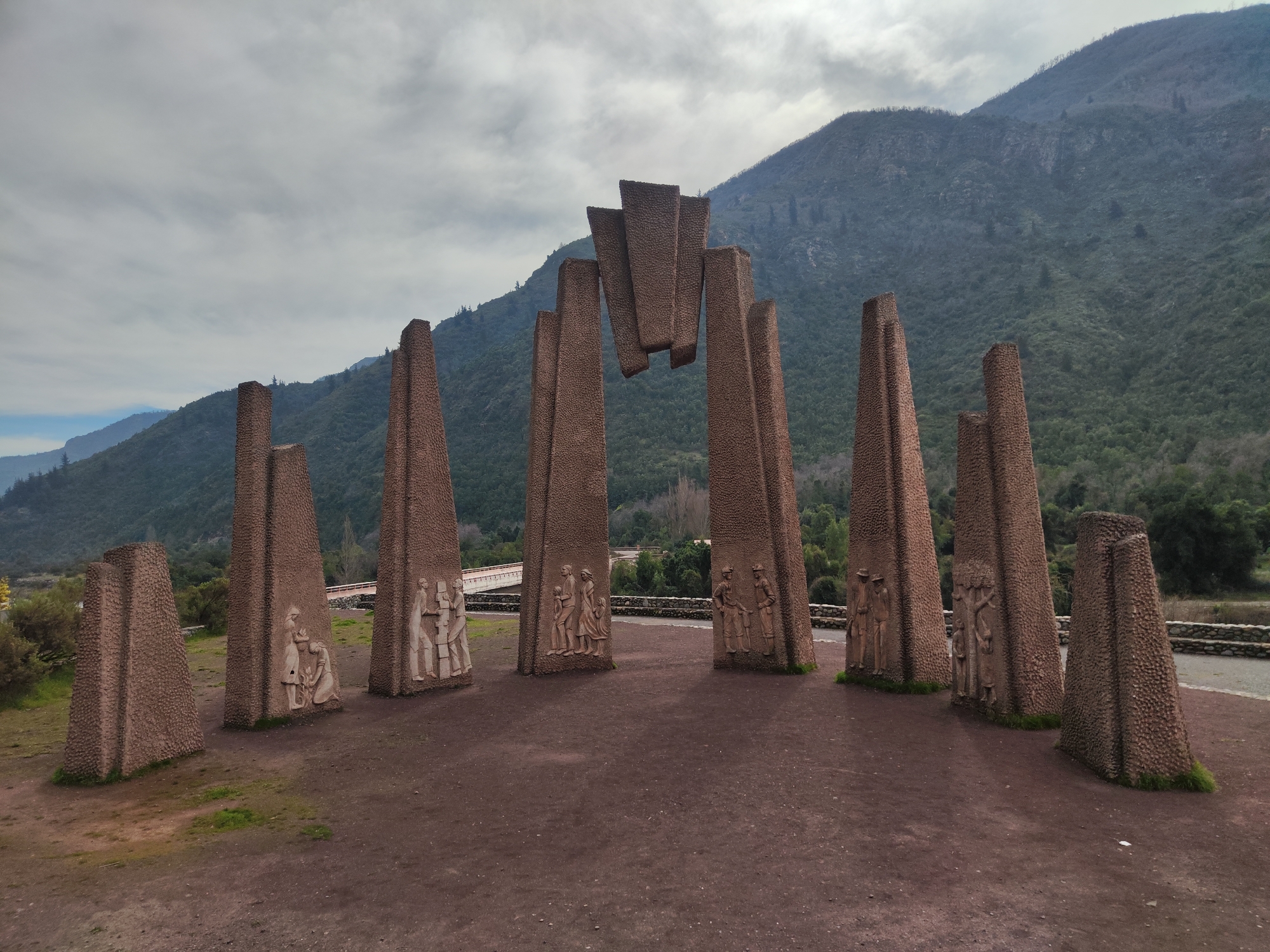 The image shows the Monumento a la Chacra in Romeral, Chile. The monument features a collection of tall, textured, triangular concrete pillars arranged in a semi-circle. Some of the pillars are adorned with bas-relief sculptures depicting groups of people in various poses, suggesting narrative scenes. In the center, several pillars lean towards each other, with flat slabs forming an arch-like structure above. The ground in front of the monument is a reddish-brown dirt path. In the background, a lush green hillside rises, leading to a mountainous landscape under a cloudy sky. A stone wall and a road are visible behind the monument, with some trees and vegetation scattered throughout the scene.
