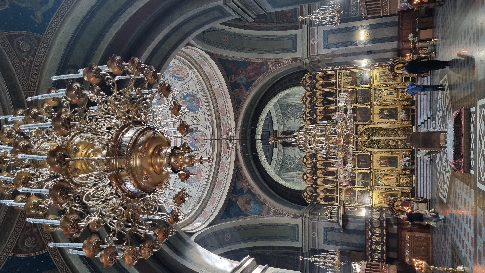 This is a full-length, vertical, eye-level shot of the interior of an Orthodox church, likely Chernivtsi Cathedral in Chernivtsi, Ukraine. The camera is positioned in the nave, looking towards the iconostasis.
Dominating the upper portion of the frame is a large, ornate, multi-tiered brass chandelier with numerous candle holders. Below the chandelier, and extending across the width of the frame, is the richly decorated iconostasis, a wall of icons and religious imagery. It is gilded and features numerous painted icons in rectangular panels, with large central doors. A large crucifix with two figures is positioned above the iconostasis, illuminated by the chandelier.
The ceiling and upper walls of the church are adorned with frescoes depicting religious scenes and figures. Arched openings reveal further painted details.
In the foreground, the floor is tiled in a geometric pattern. Several people are present: a woman in a dark top and light pants walks from the left towards the center; a man in a blue shirt and dark pants walks towards the right; and a person in a black robe walks away from the camera towards the right. There are also several large brass candle stands with lit candles visible on the left and right sides of the frame, as well as floral arrangements in front of the iconostasis. The overall impression is one of grandeur and religious solemnity.